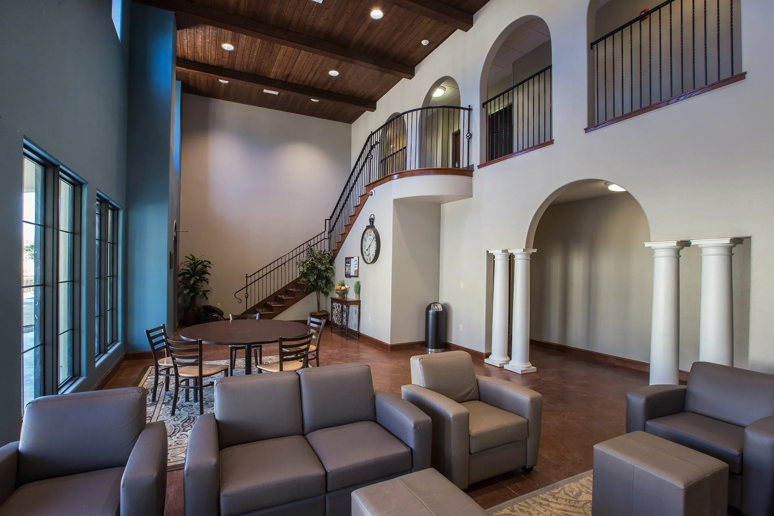 Interior of a spacious lobby with a high wooden ceiling, large windows, gray furniture, a round table with chairs, a staircase, columns, and decor.