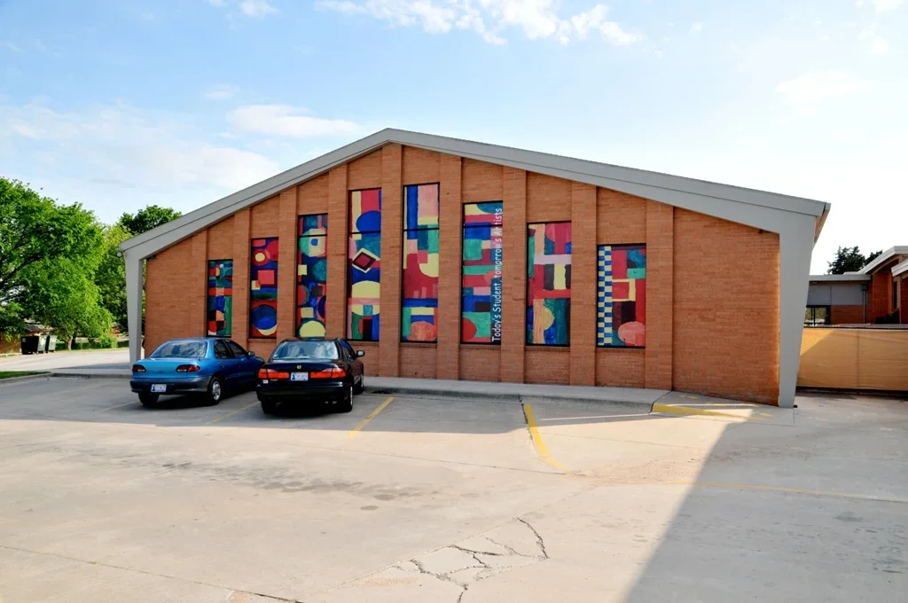 Front view of a brick building with a sloped roof, decorated with colorful abstract art panels in vertical frames. Three cars are parked in front of the building, and there are parking lot lines and a few trees in the background.