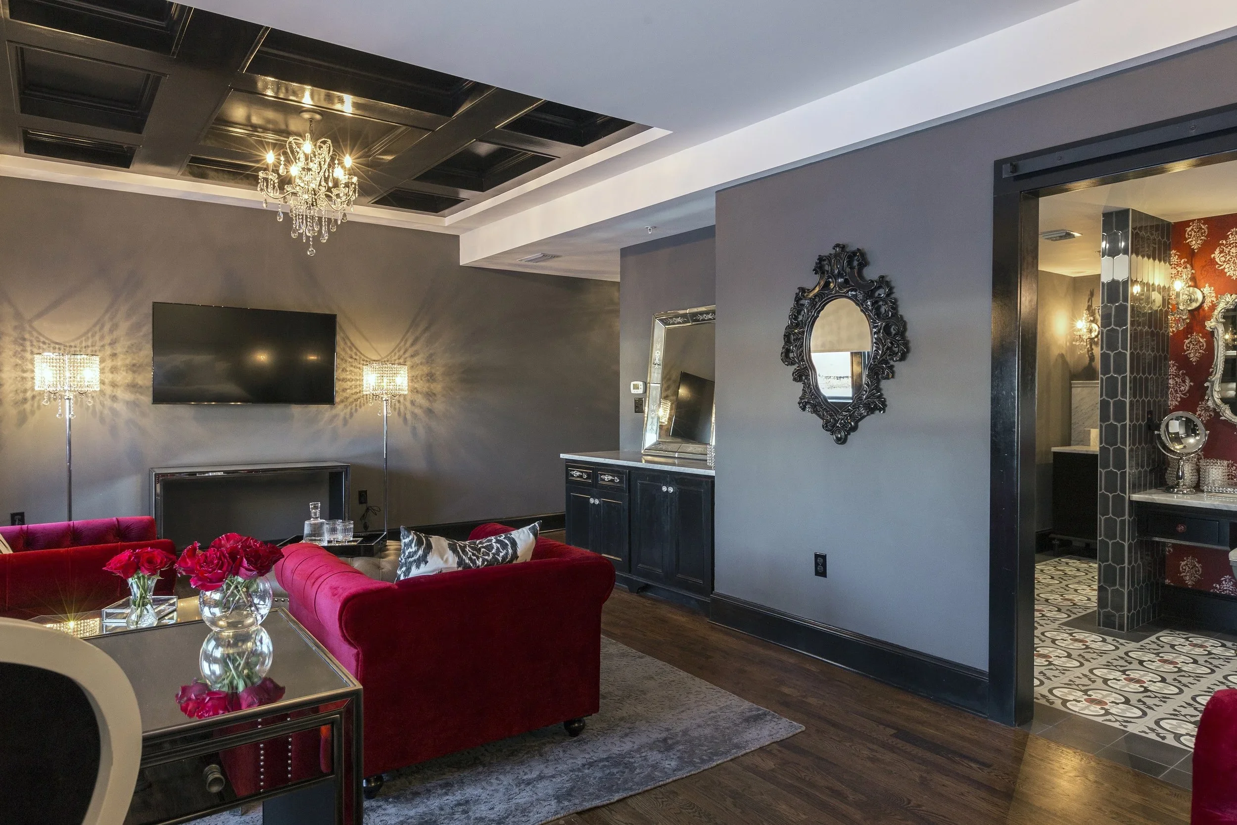 Luxurious living room with gray walls, a chandelier, a flat-screen TV, two floor lamps, red velvet sofas, decorative pillows, a glass coffee table with flowers, a black cabinet with a mirror, and a doorway leading to a patterned bathroom with black, 