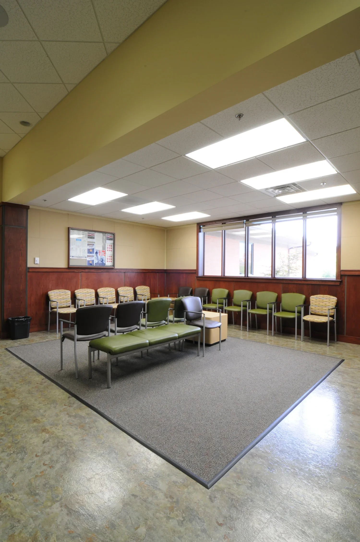 Empty waiting room with green and patterned chairs, a small table, and large windows letting in natural light.