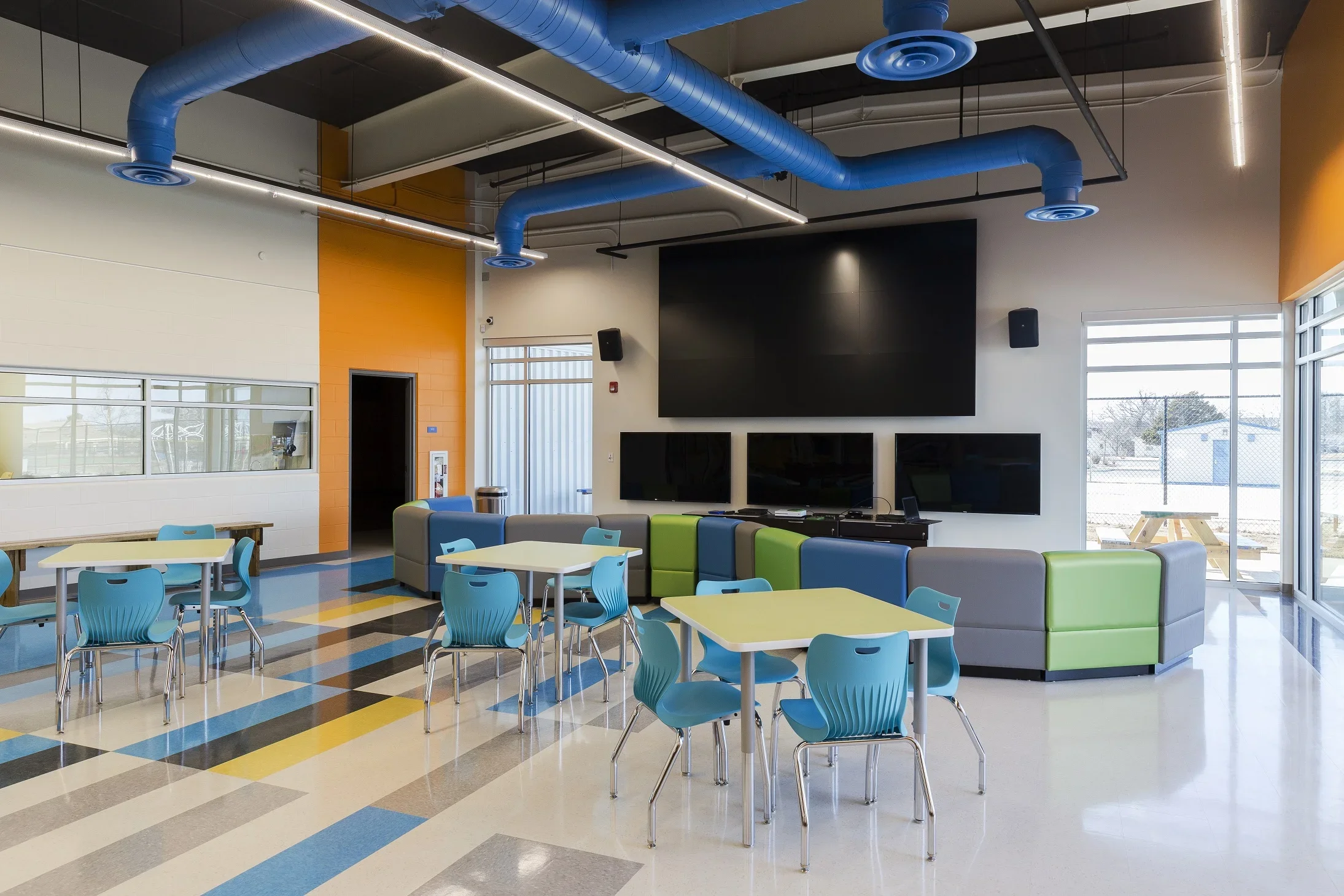 Colorful lounge area with blue chairs, white tables, and a gray and green cushioned seating area. Large TV screens mounted on the wall and a big flat screen TV. Bright natural light through windows on the right and a door on the left. Industrial ceil