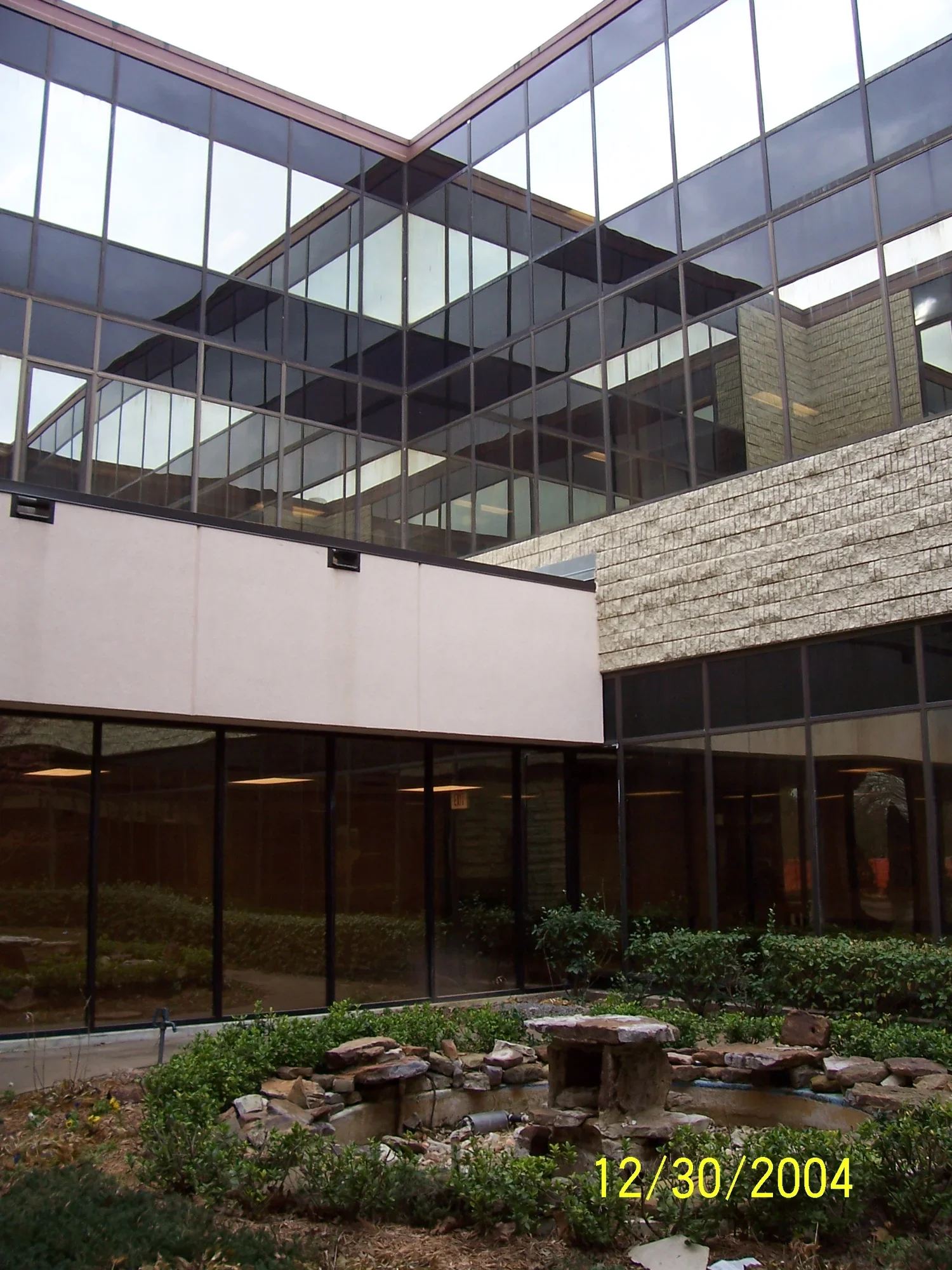 Photo of a modern glass building with multiple levels, reflecting the sky, and a small garden with rocks and greenery in front.