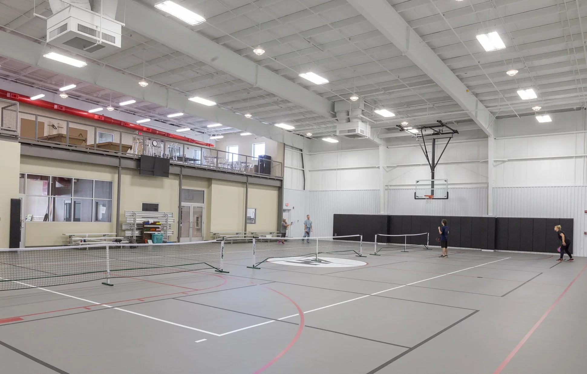 Indoor gymnasium with a basketball hoop, tennis nets, and four children playing badminton. There are upper storage shelves with chairs and boxes, and the gym has bright lighting and a high ceiling.