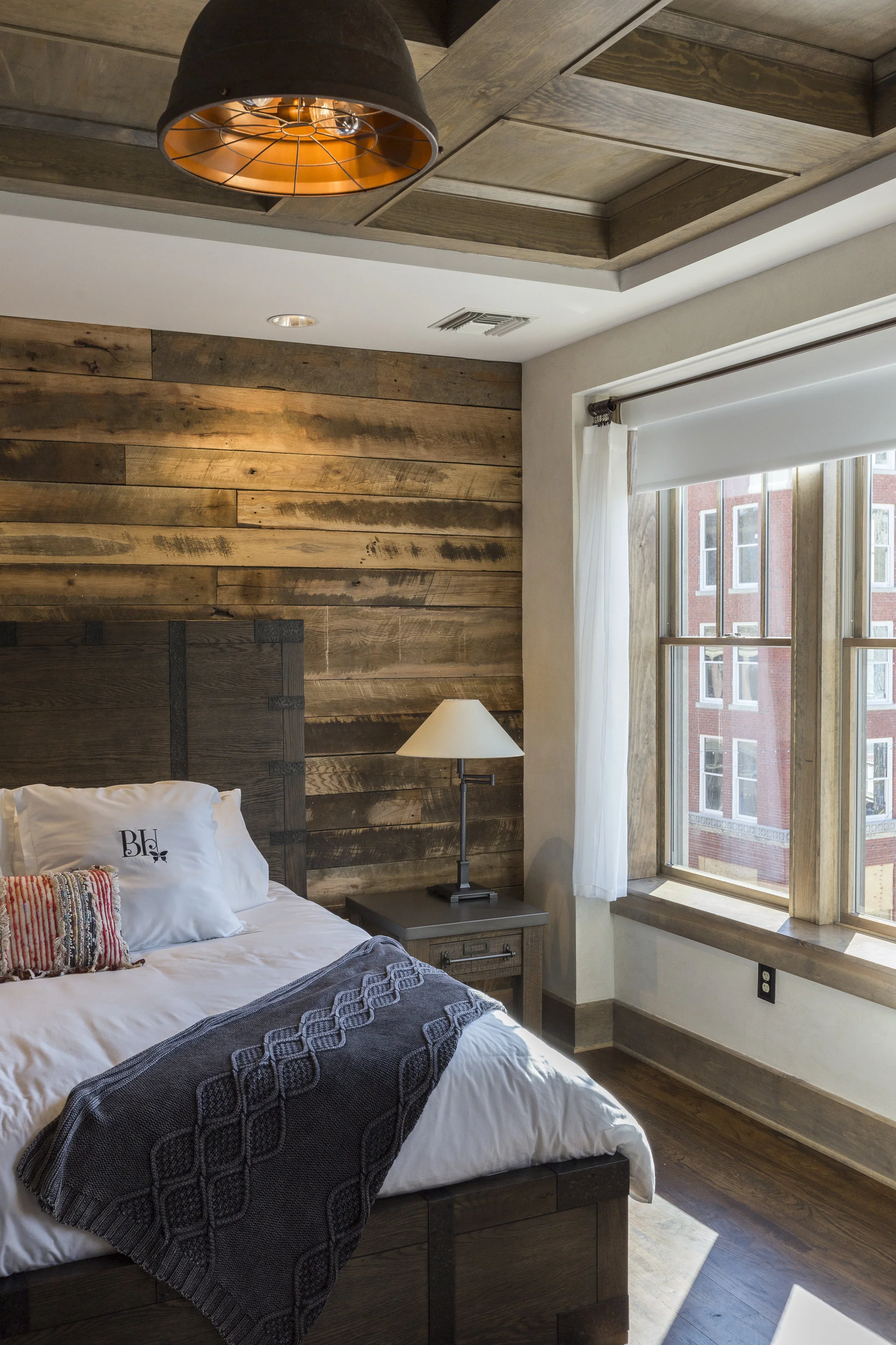 A bedroom with wood-paneled walls, a bed with white bedding, a decorative pillow, and a gray blanket. There is a bedside table with a lamp, and large windows with white curtains letting in natural light.