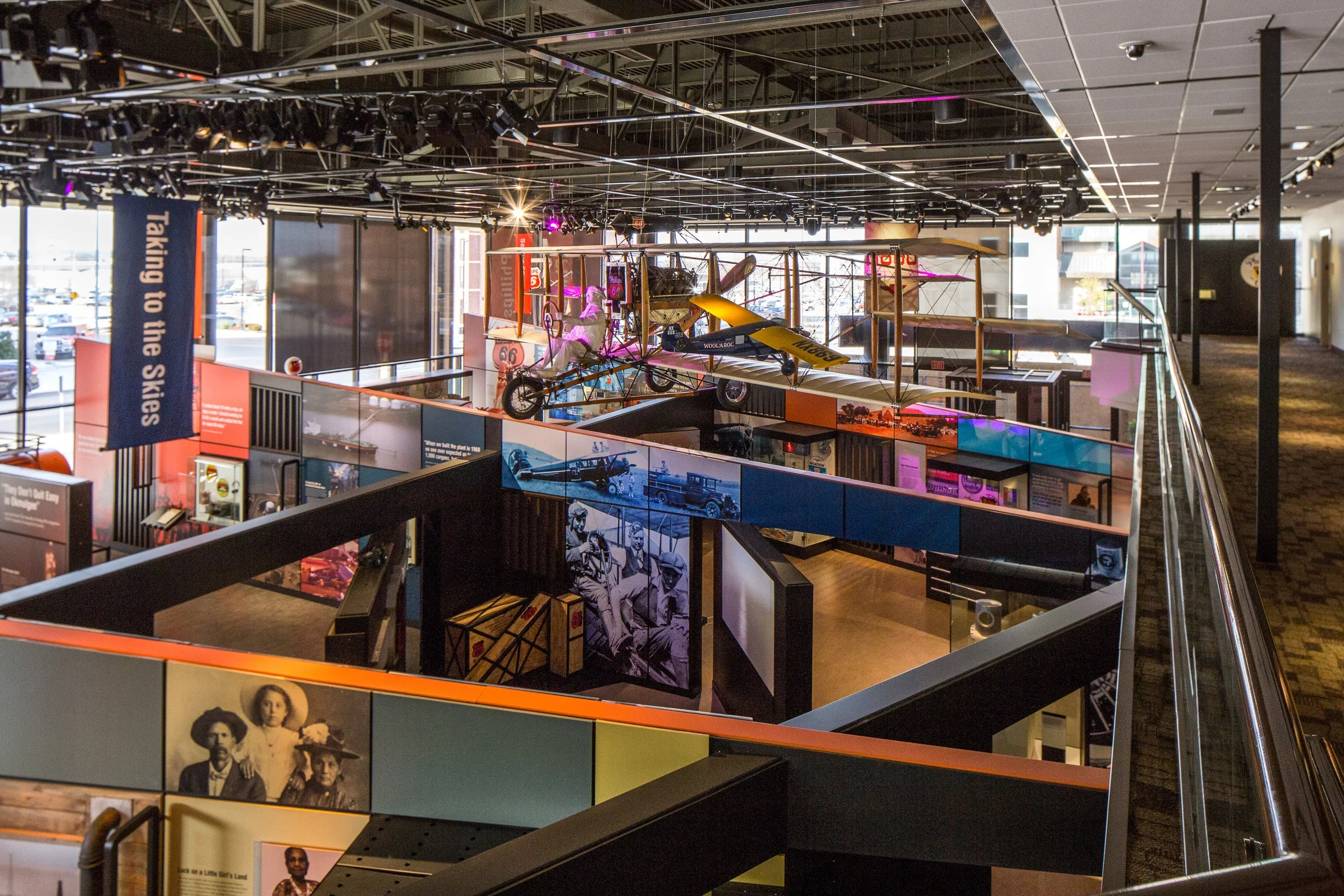 Interior of a museum exhibit showcasing vintage aircraft, motorcycles, and historical displays, with large windows, banners, and informational panels.