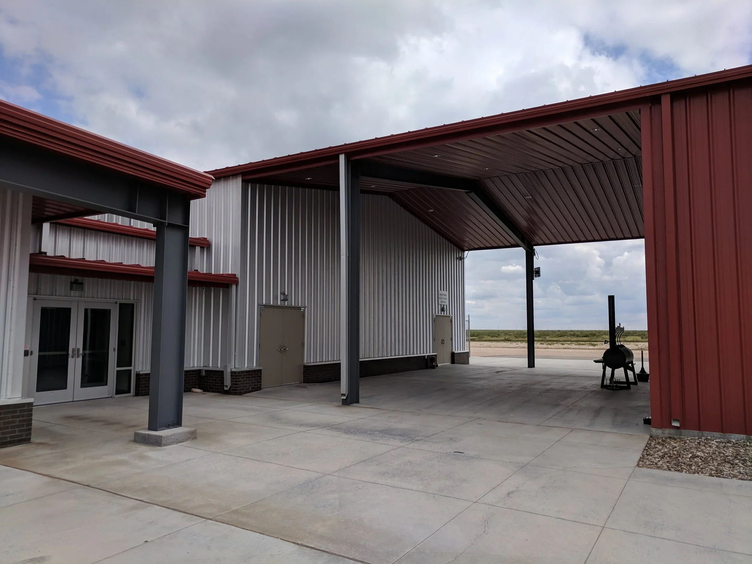 Empty outdoor covered area with metal building walls, a barbecue smoker, and an open landscape with cloudy sky in the background.