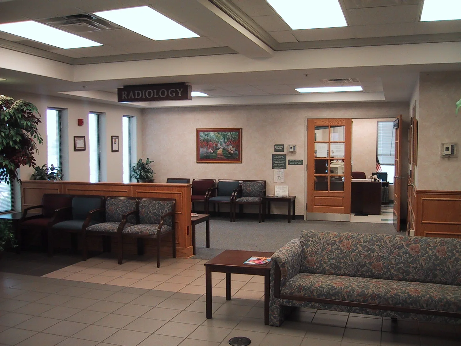 Interior of a medical facility waiting room with chairs, a couch, potted plants, and a sign that reads 'Radiology' hanging from the ceiling.