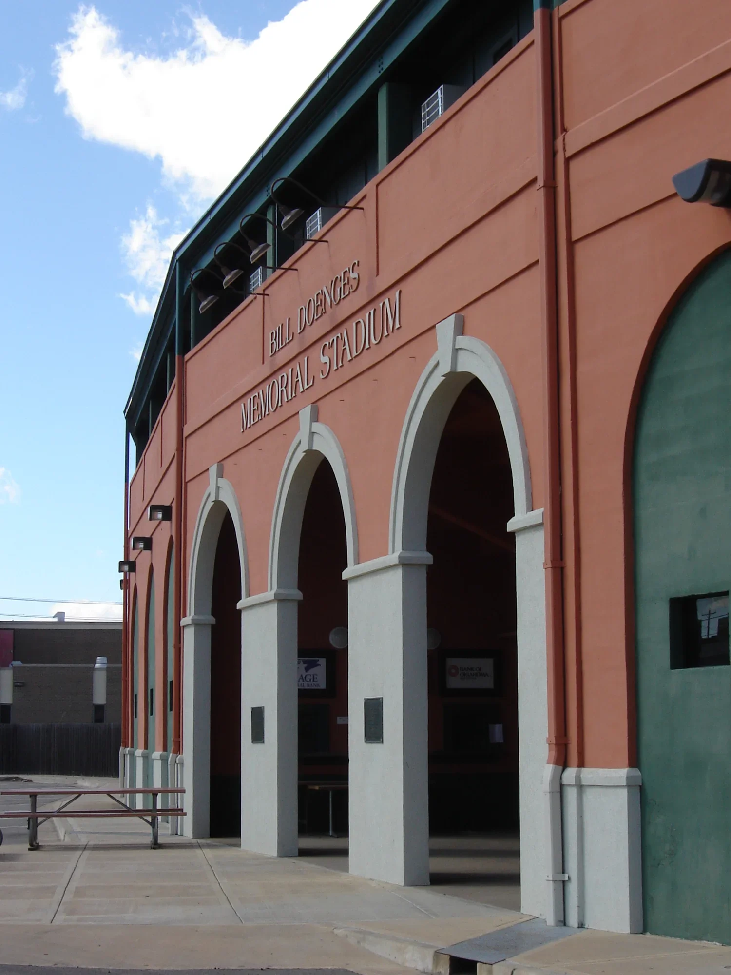 Exterior of Bill Doenges Memorial Stadium with three arched entryways and signage, against a blue sky with clouds.