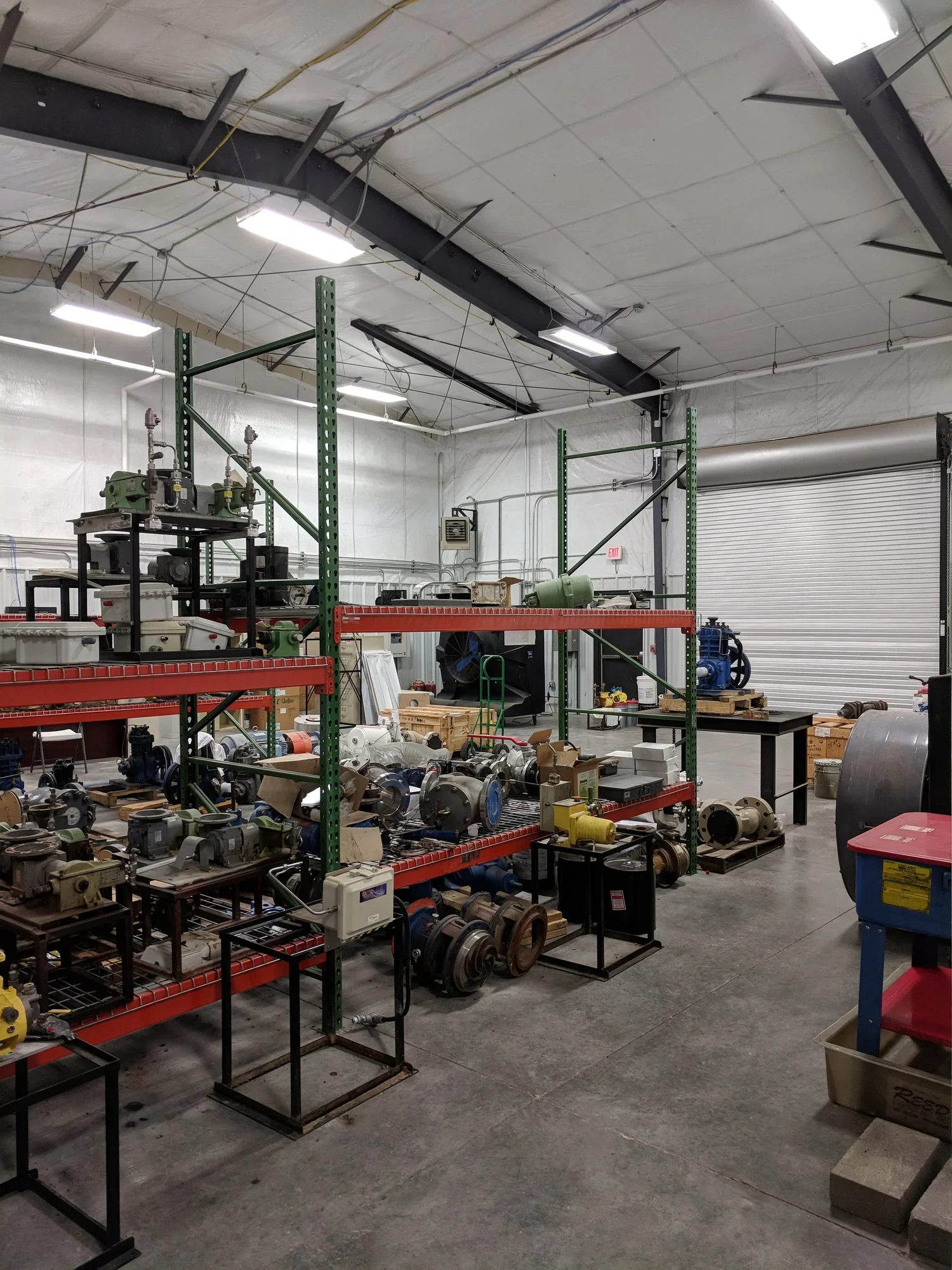 Industrial workshop with metal parts and machinery on shelves and tables, and a roll-up garage door in the background.