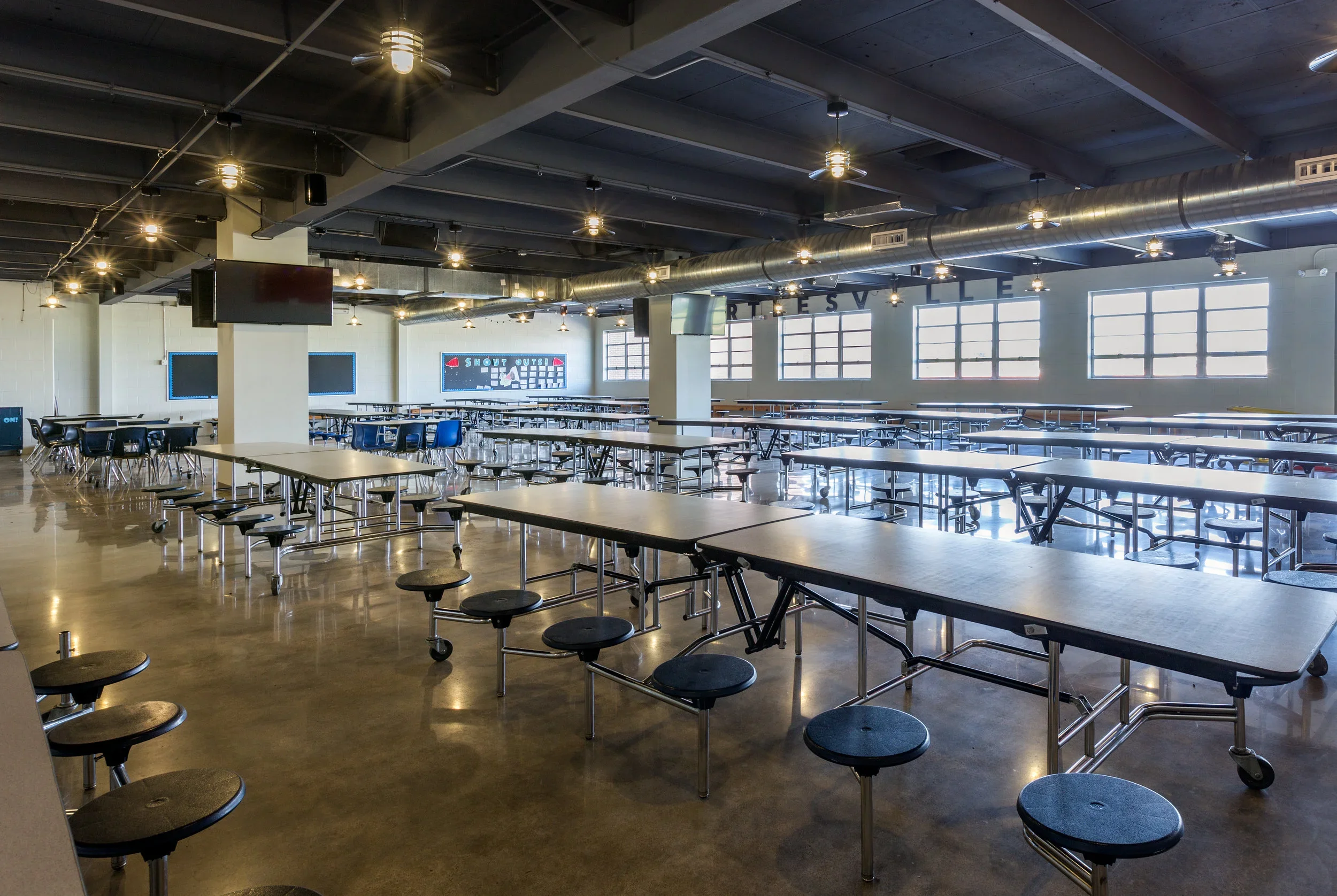 Empty cafeteria with long tables, black stools, and large windows letting in natural light. Industrial ceiling with exposed ductwork and hanging lights.