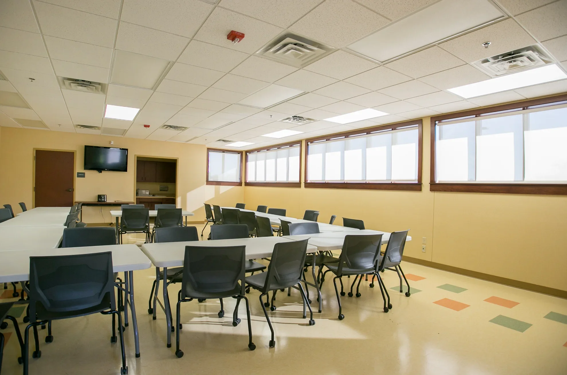 Empty conference room with tables and chairs, a wall-mounted TV, and windows letting in natural light.