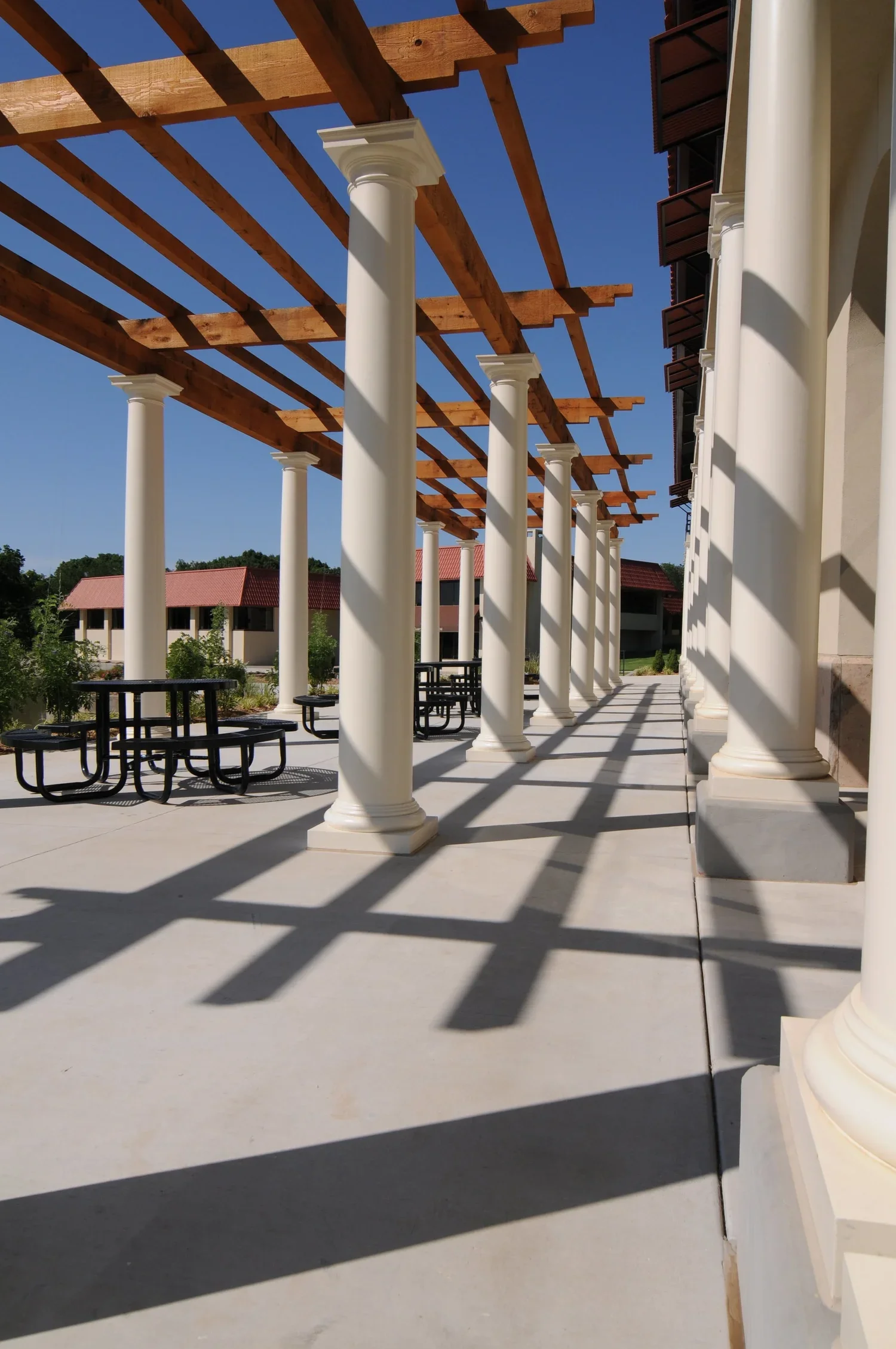 Columns supporting a wooden pergola on a sunny patio with picnic tables, cast shadows, and a building with red roofs in the background.