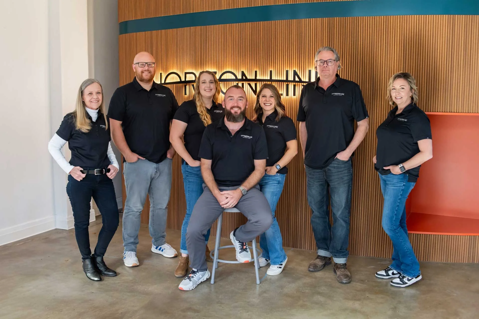 Group of the Horizon Line team standing in front of a wooden wall with a illuminated sign that reads 'HORIZON LINE', all wearing black shirts with a logo, smiling for the photo.