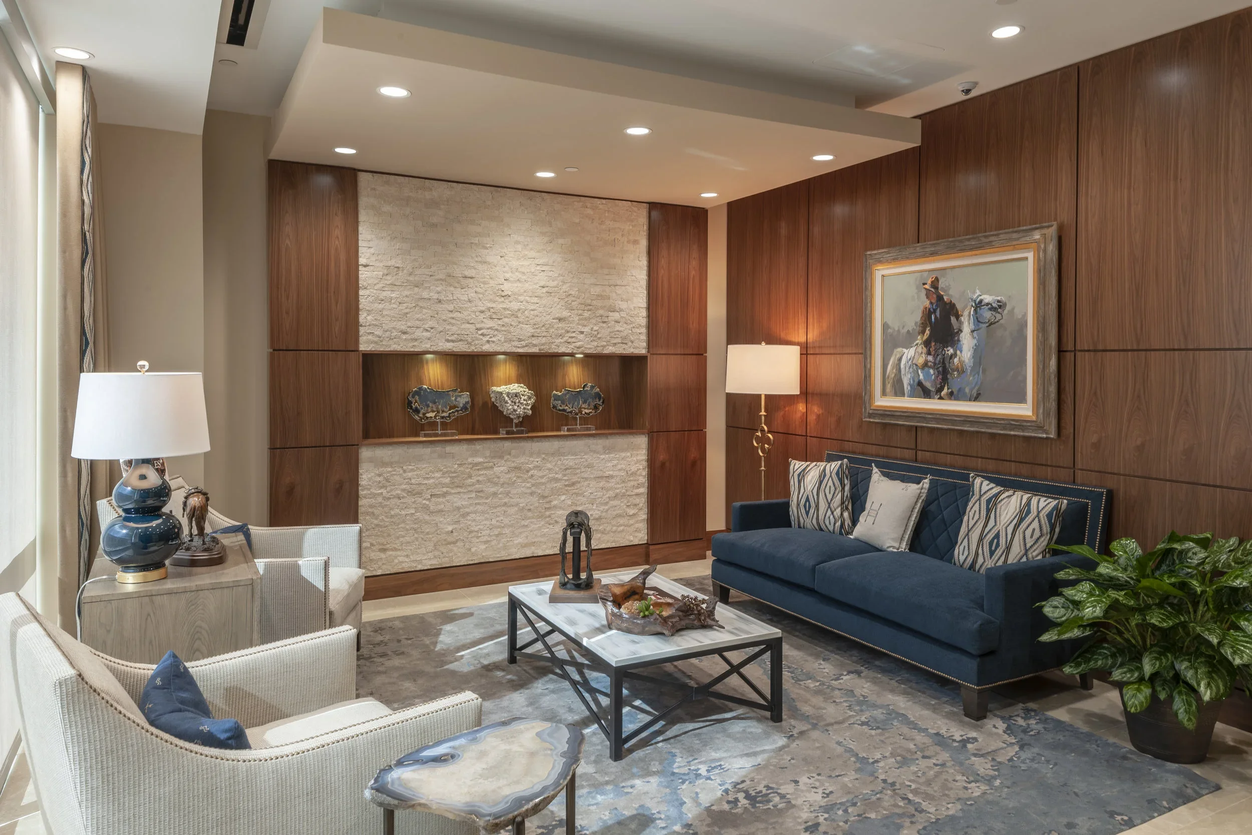 A modern office lobby with beige and wooden walls, a blue velvet sofa with patterned and gray pillows, a white armchair, a coffee table, and decorative art pieces on the wall and shelf, with a large area rug and potted plant.