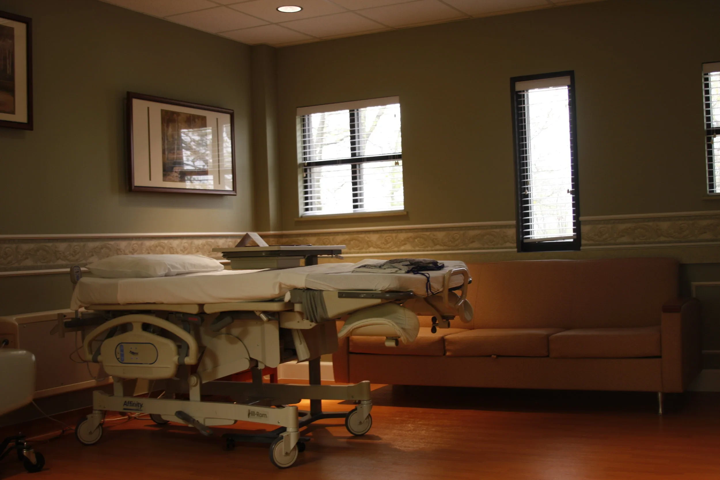 Empty hospital room with a bed and a couch, windows with blinds, and framed pictures on the wall.