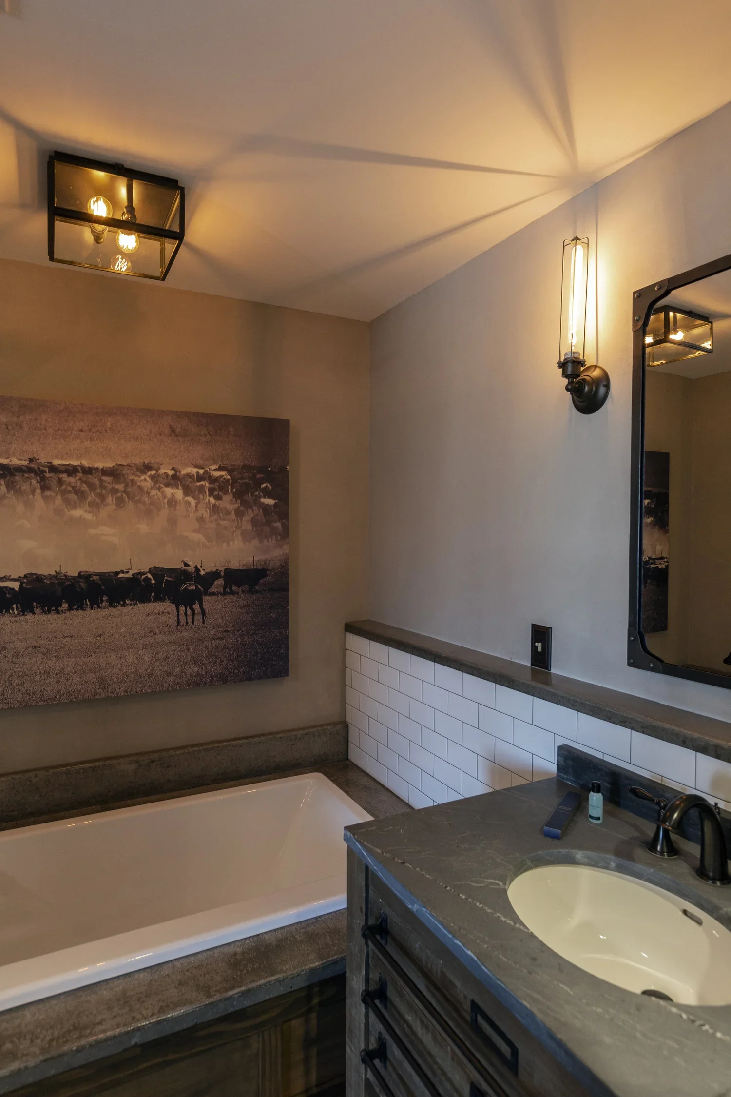 Modern bathroom with a white bathtub, gray marble countertop with an oval sink, and a large mirror. The wall has white subway tiles, and a black wall-mounted light fixture. A large black and white photograph of cattle and a herd of buffalo decorates 