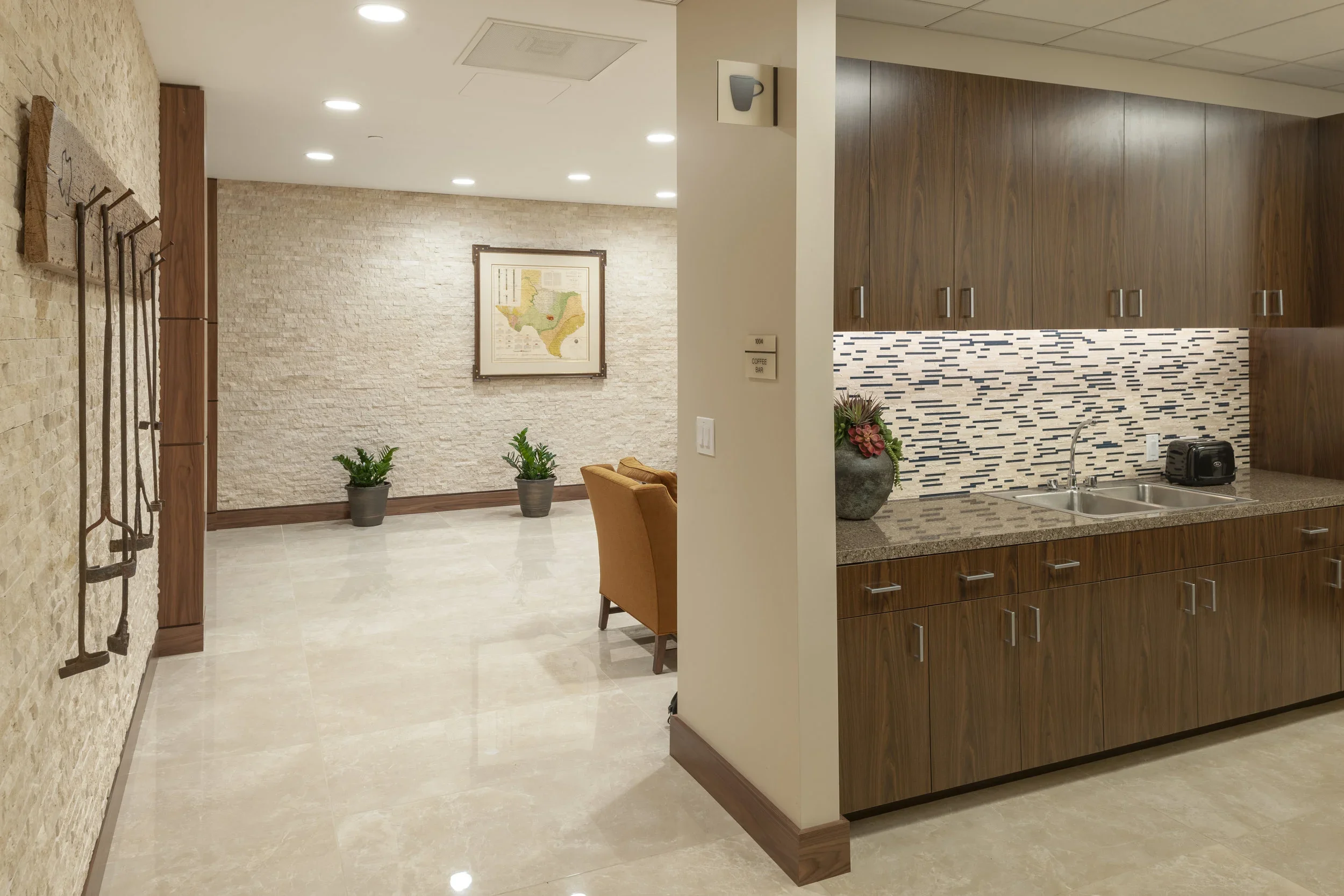 Interior of a modern office or lobby area with a stone wall, potted plants, a framed picture of Texas, and a kitchenette with wooden cabinets, a mosaic tile backsplash, a sink, and a coffee maker.