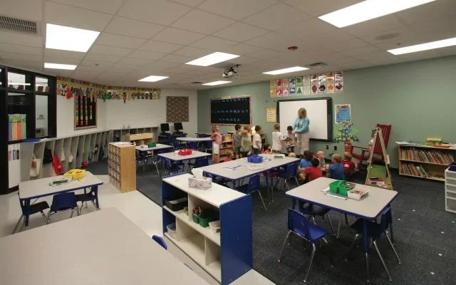 Classroom with children and a teacher at the whiteboard, desks with blue chairs, and educational decorations.