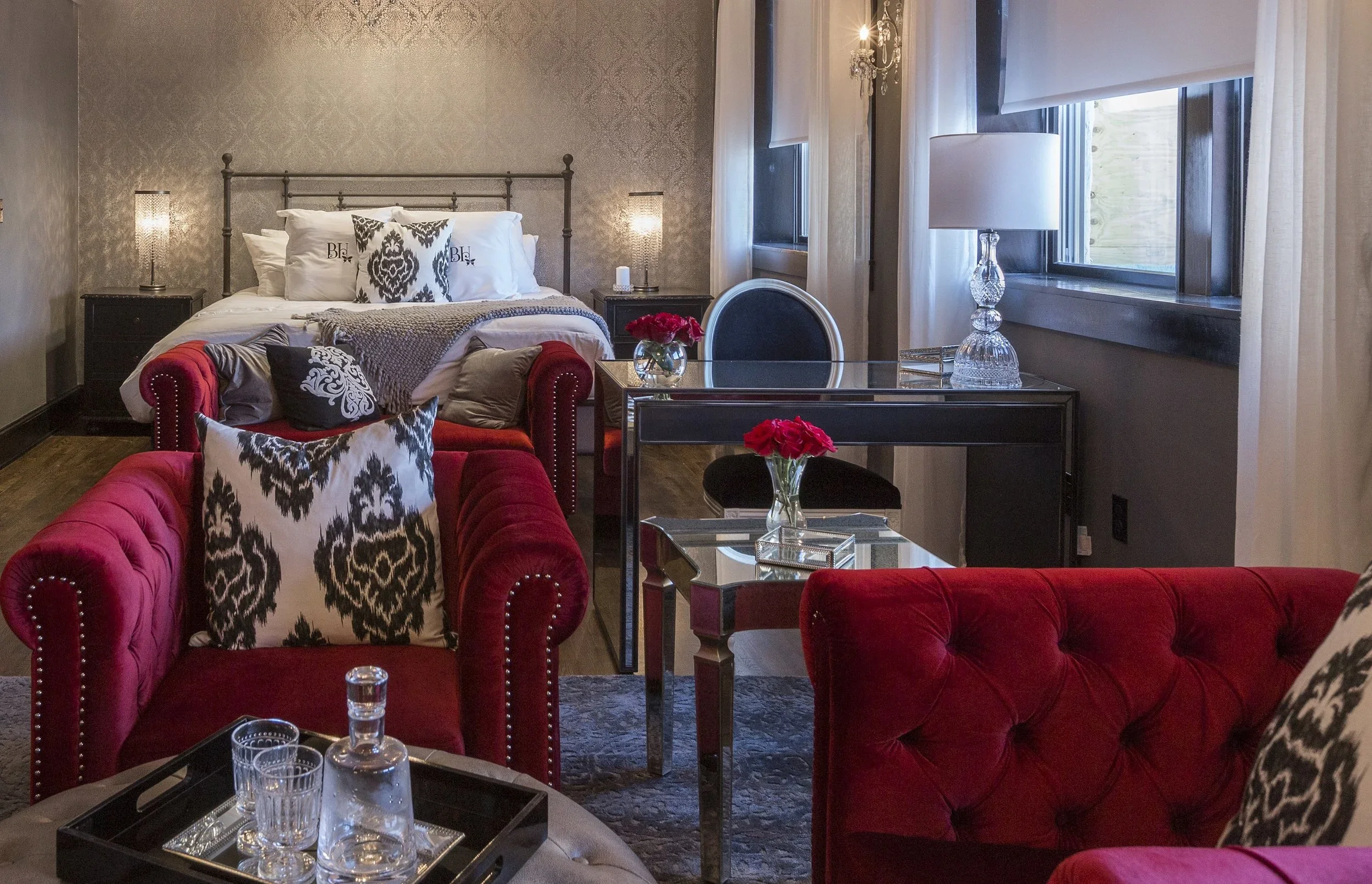 A cozy hotel room with a bed, two matching bedside tables with lamps, a black vanity desk with a glass lamp and a vase of red flowers, a black and white upholstered chair, a red tufted sofa with patterned pillows, a glass tray with a water bottle and glasses, and a window with white curtains.