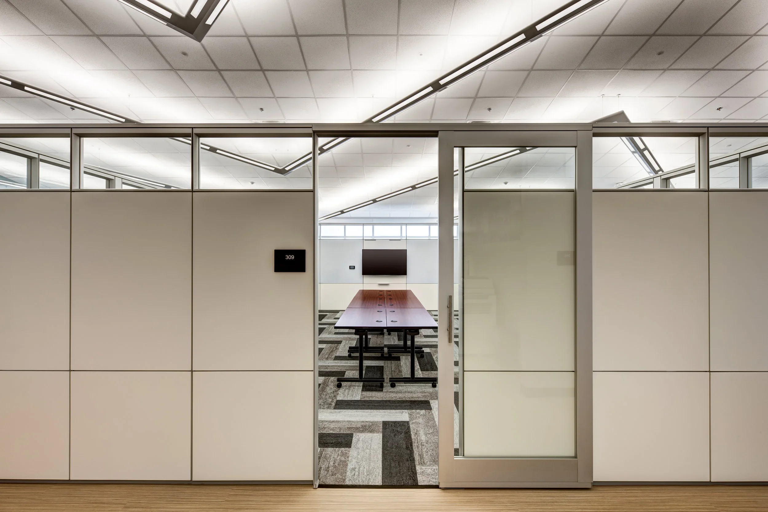 View of a modern office conference room through sliding glass doors, with a long wooden table, patterned carpet, and a large monitor on the back wall.