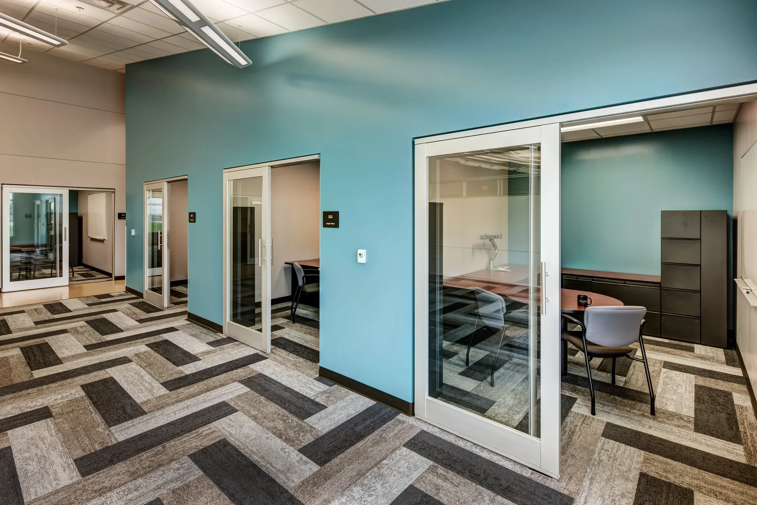 An office corridor with individual glass-door rooms, some with chairs and desks inside, and a patterned carpet floor.