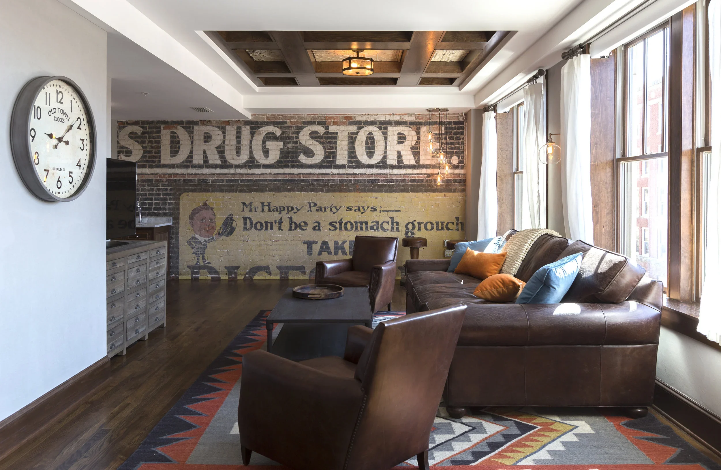 Living room with a brick wall mural that reads 'S Drug Store' and features a vintage advertisement with a boy holding a tray, a large wall clock, leather armchairs, a patterned area rug, large windows with white curtains, and a brown leather sofa with colorful pillows.