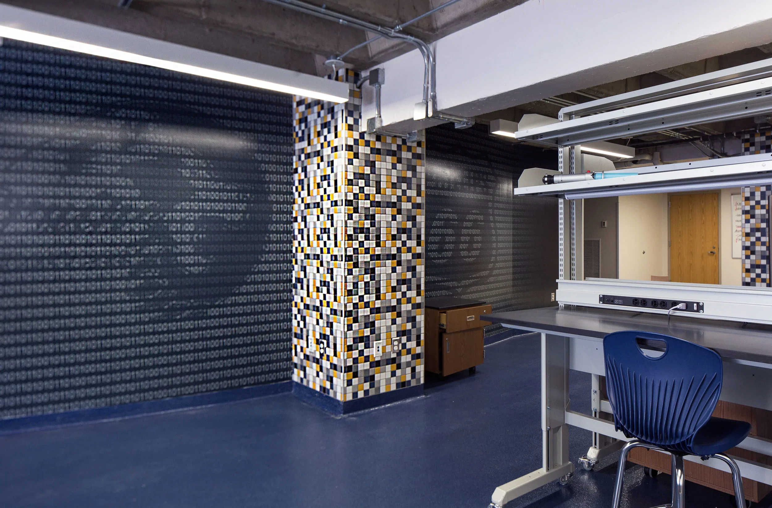 Interior of a room with a black and white checkered pillar, walls covered with binary code pattern, and a workspace with a blue chair, countertop, and shelves.