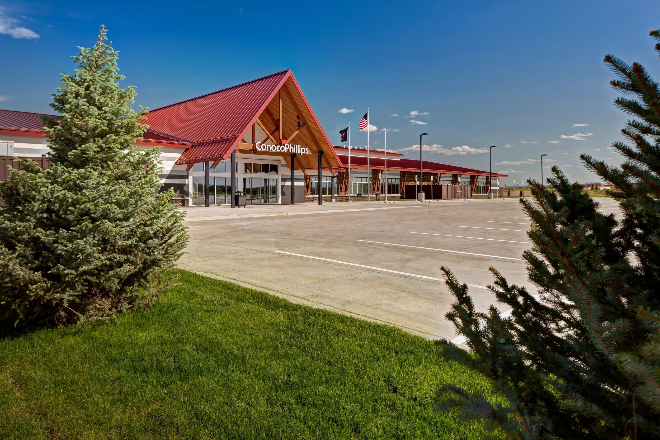Empty parking lot outside a modern building with a red roof, labeled 'ConocoPhillips,' under a blue sky with some clouds, flanked by small evergreen trees and green grass.
