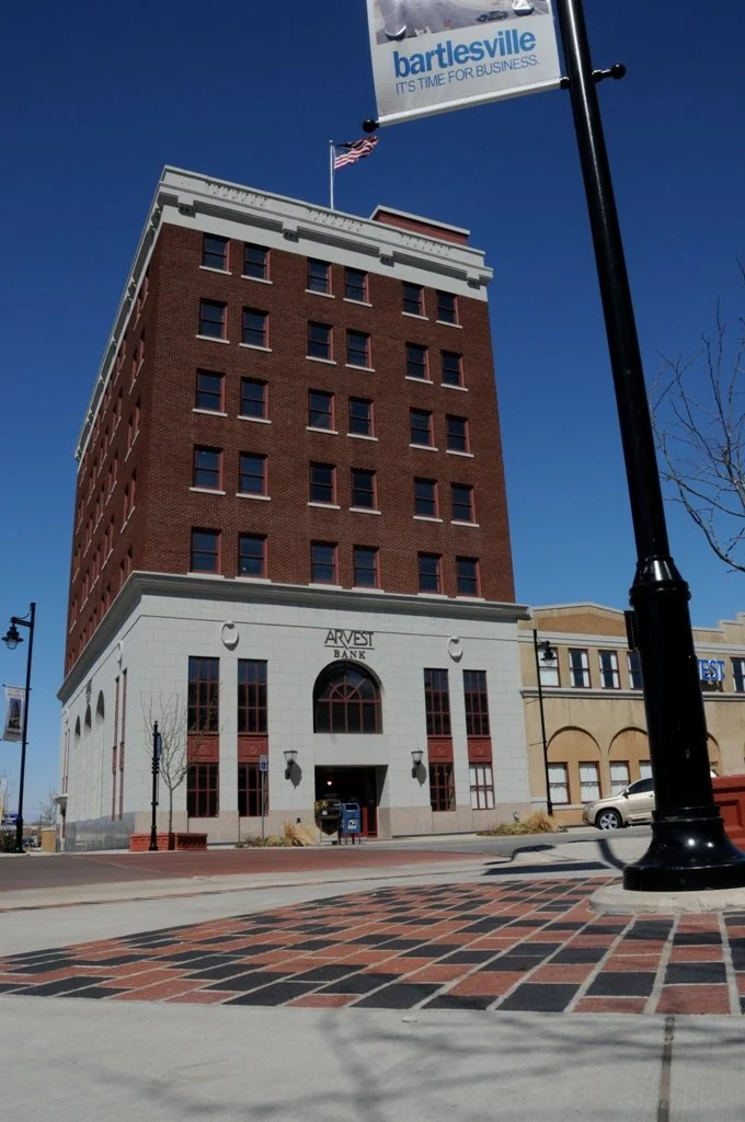 A tall brick building with the sign 'Arvest Bank' on its white base, situated on a city street with a brick and black patterned sidewalk, a lamppost, flags, and a blue sky.