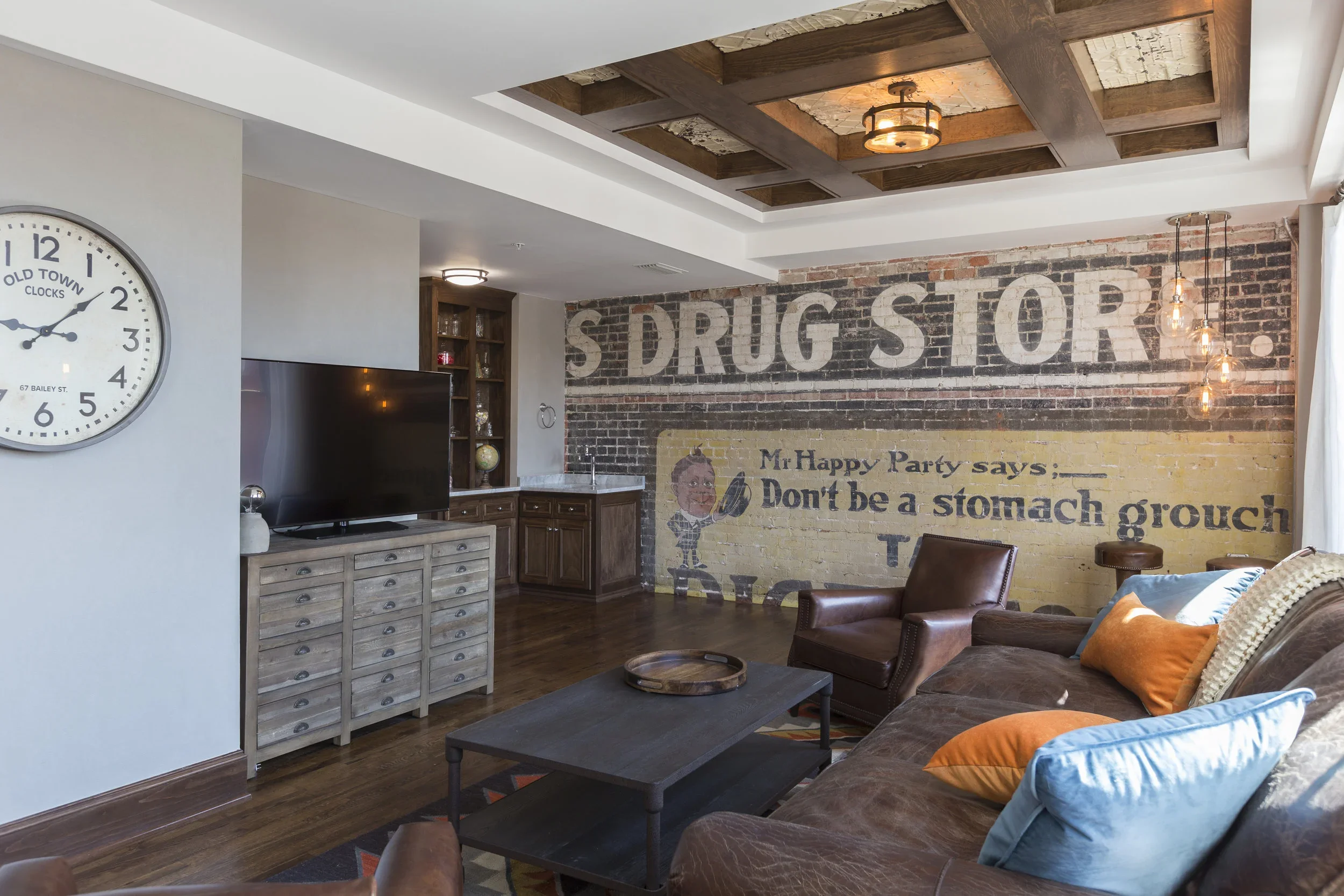 A living room with a brick wall painted with a mural of a drug store. The mural includes a cartoon character and the text: 'Mr Happy Party says: Don't be a stomach grouch.' The room has a brown leather sofa with colorful pillows, a wooden coffee tabl