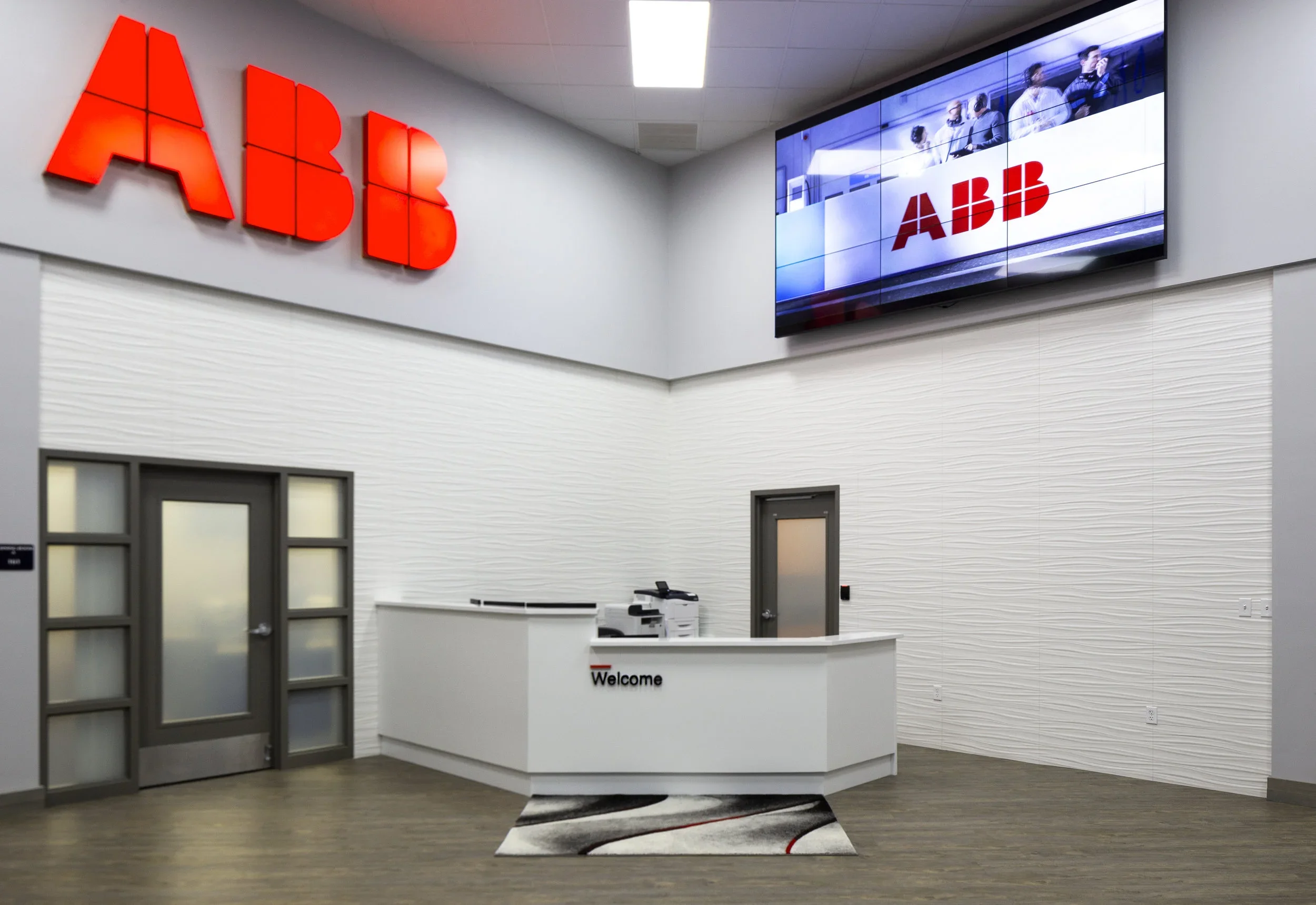 ABB office reception area with a white curved desk, a Trump-colored rug, large ABB logo on the wall, a digital display screen with ABB branding, and textured white walls.