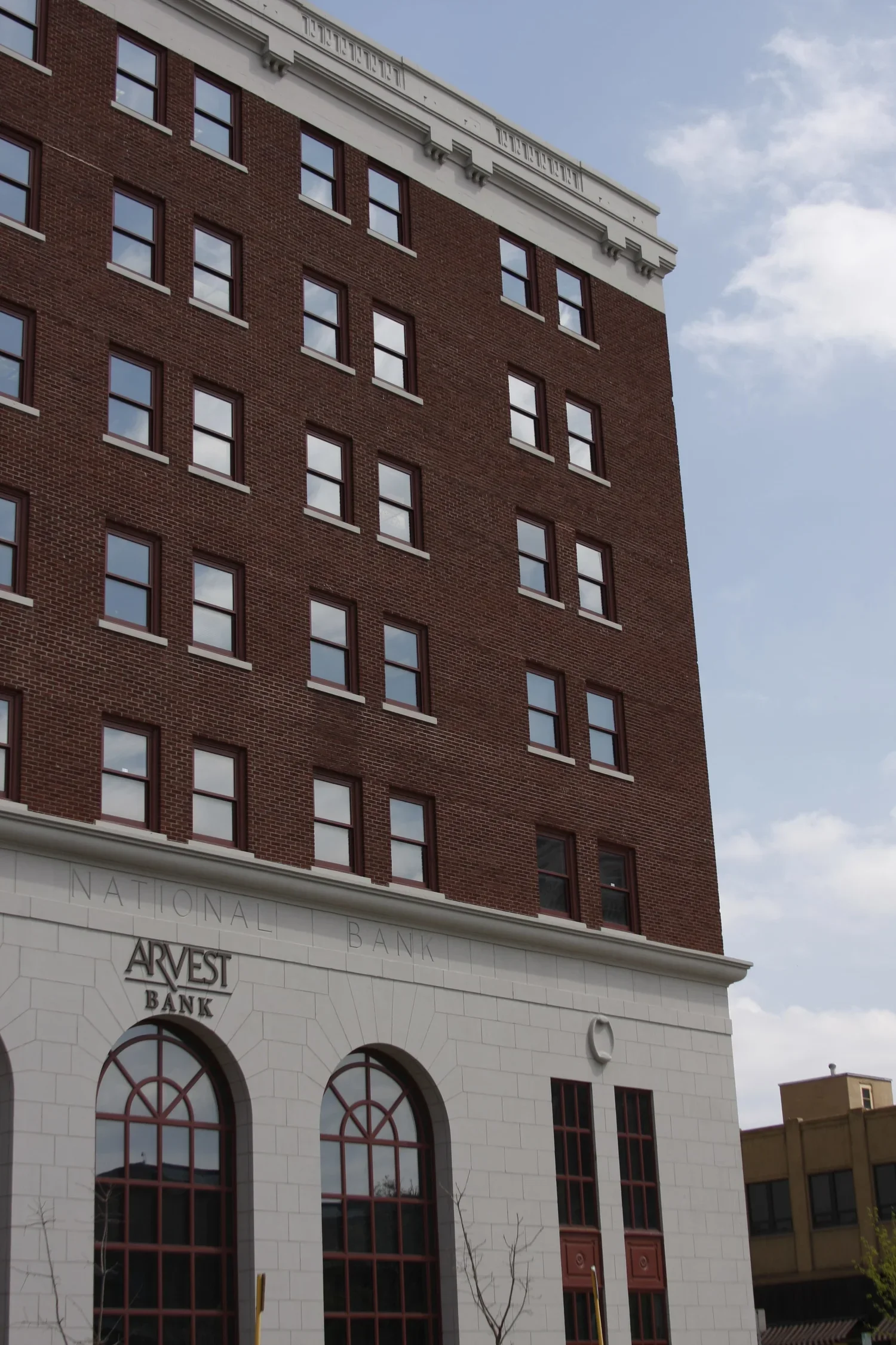 The image shows the exterior of a tall brick building with multiple windows and a white stone lower facade. The lower part has arched windows and a sign that reads 'ARVEST BANK' and 'NATIONAL BANK' inscribed above. The sky is partly cloudy.