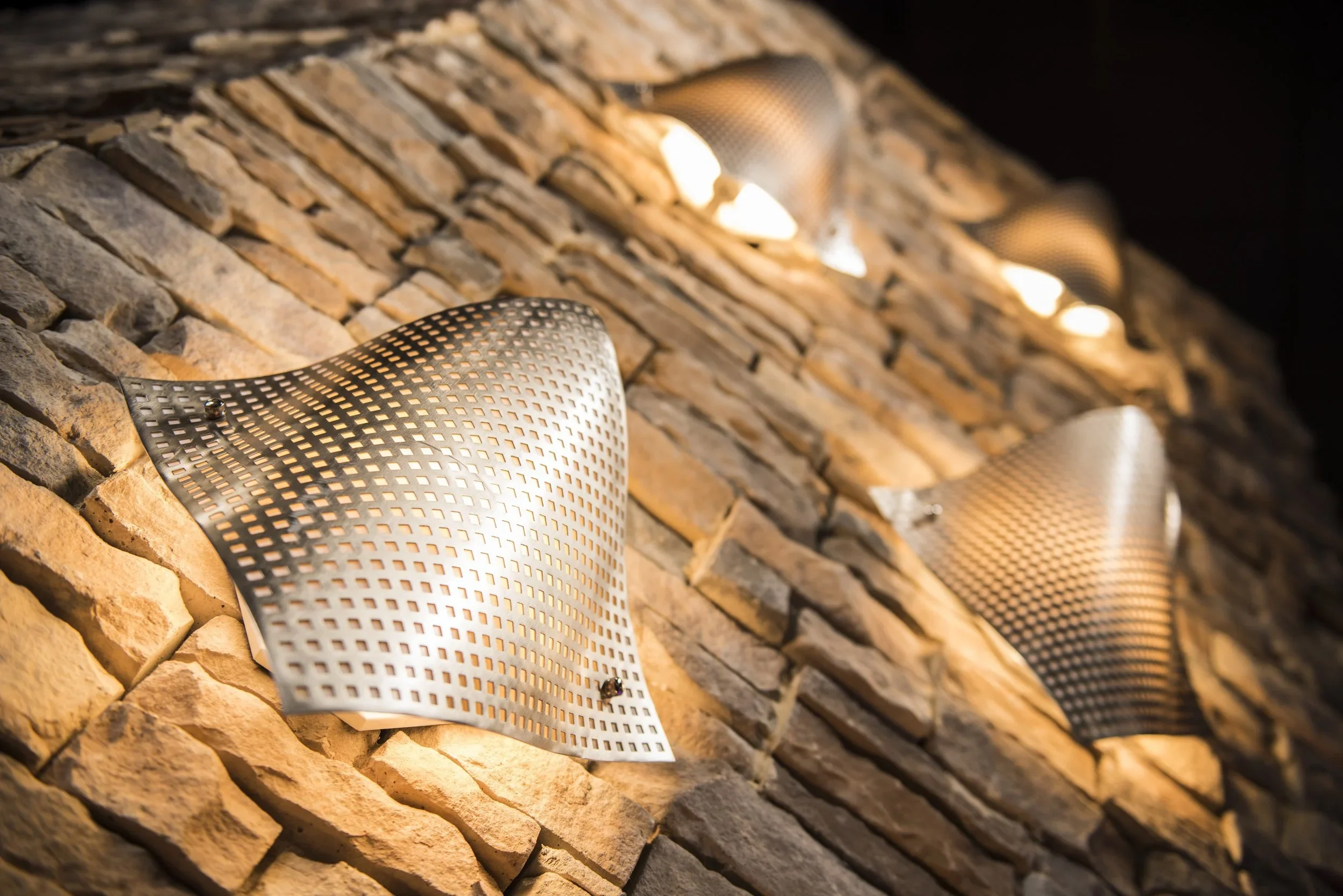 Close-up of sleek, modern wall-mounted lights with perforated metal covers, illuminating a textured stone wall.