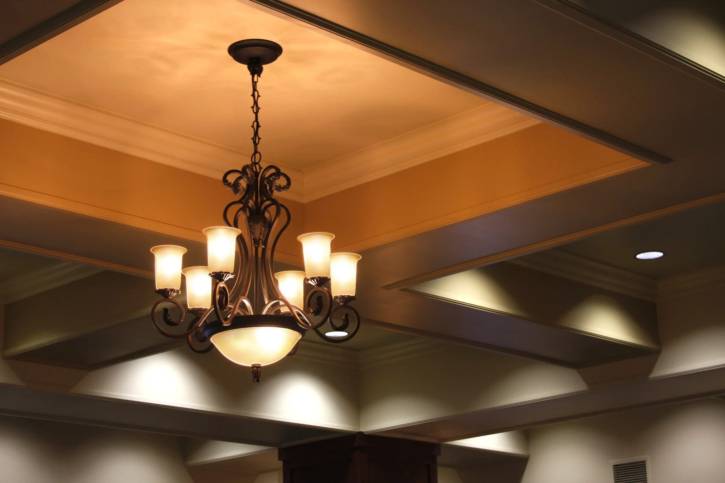 An ornate chandelier with multiple lights hanging from a ceiling with recessed lighting and crown molding in a warm, decorated interior space.