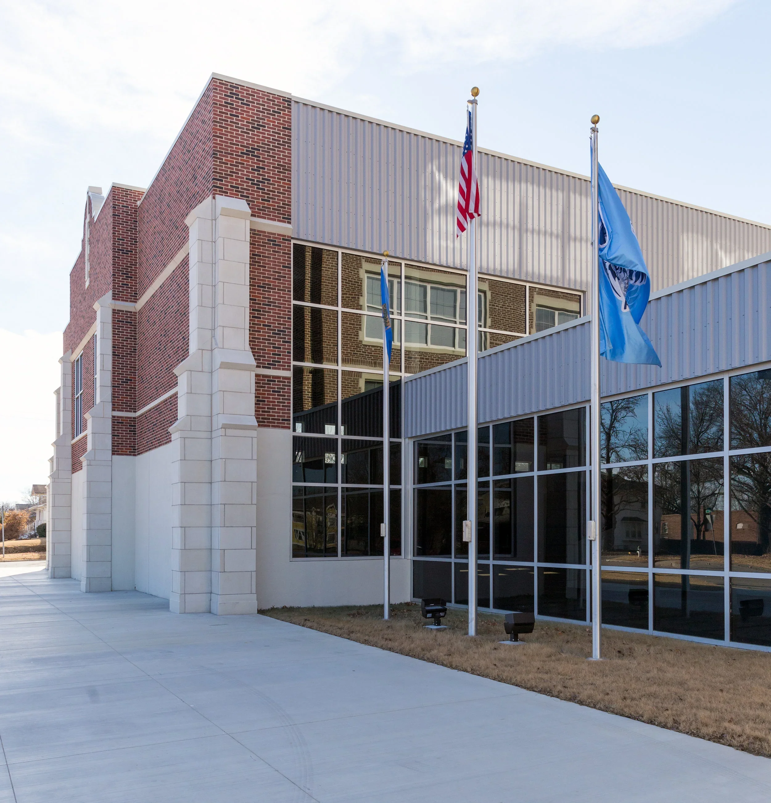 Exterior view of a modern Bartlesville Public School building with brick and glass facade, three flagpoles with American, Oklahoma, and another flag, trees reflected in windows, and a paved sidewalk.