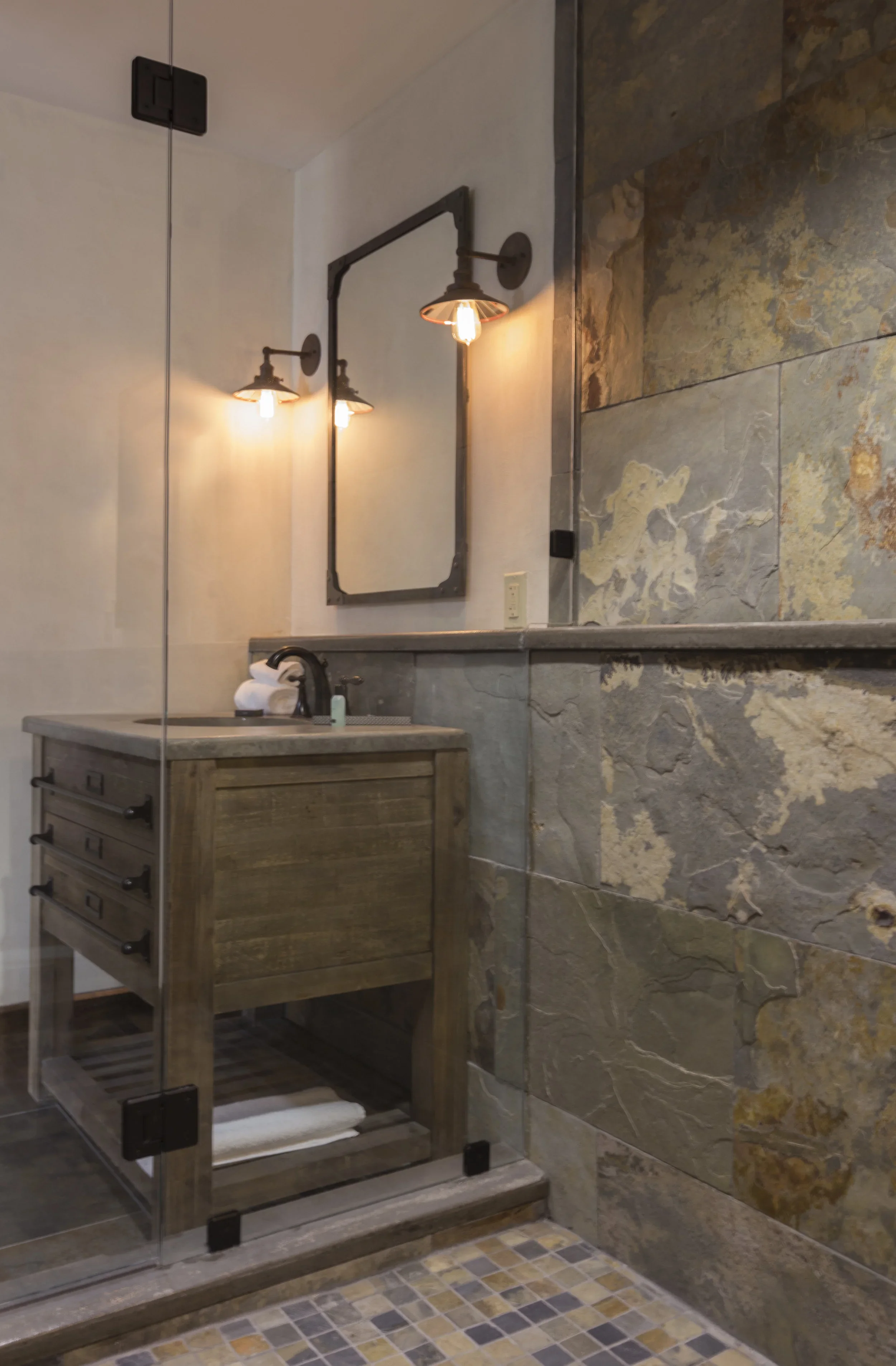 A rustic bathroom with stone and tile walls, a wooden vanity with a black faucet, wall-mounted industrial-style lights, and a glass shower enclosure.
