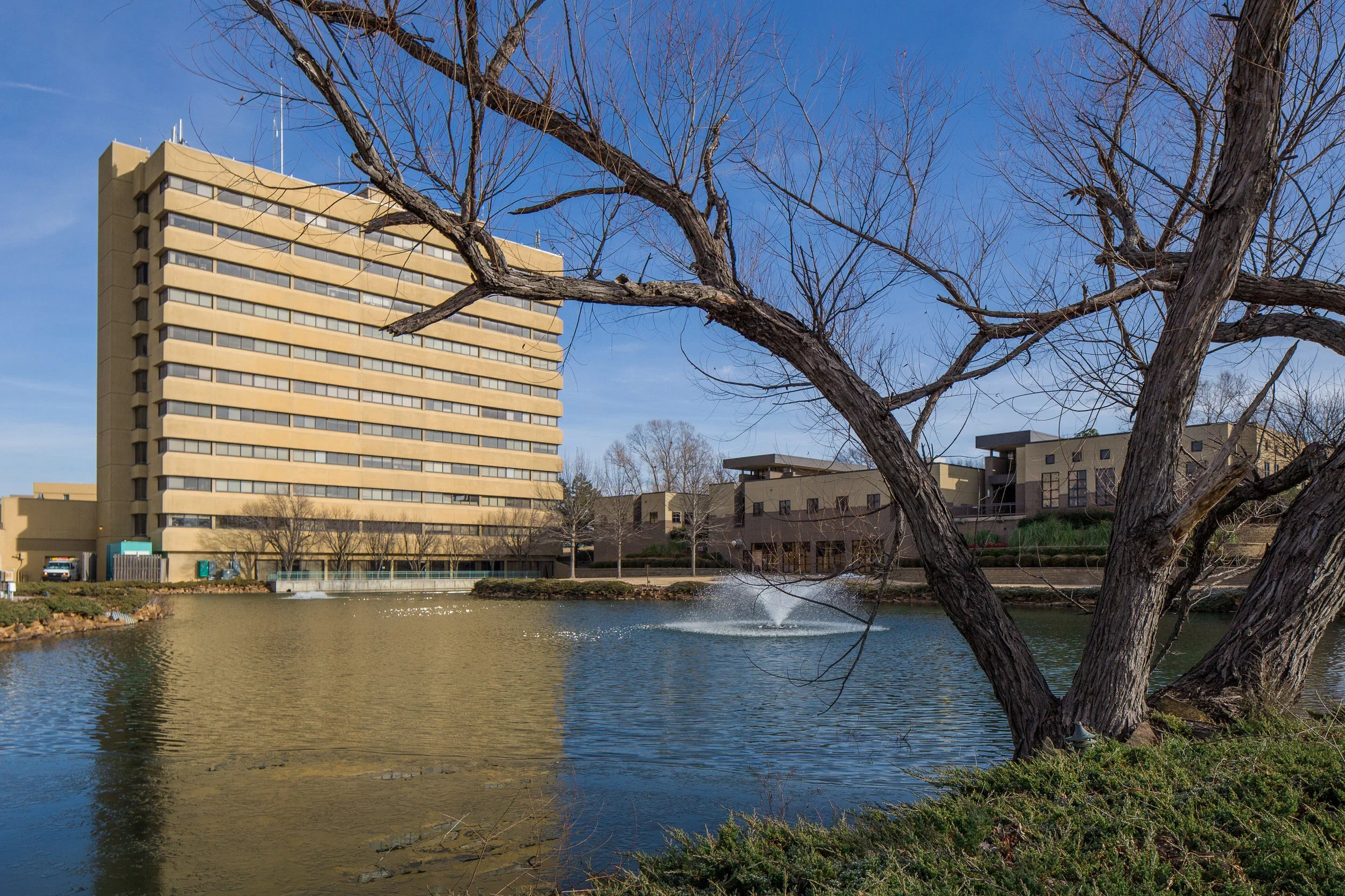 Exterior of Jane Phillips Surgical Hospital next to a pond with a fountain, trees, and a clear blue sky.