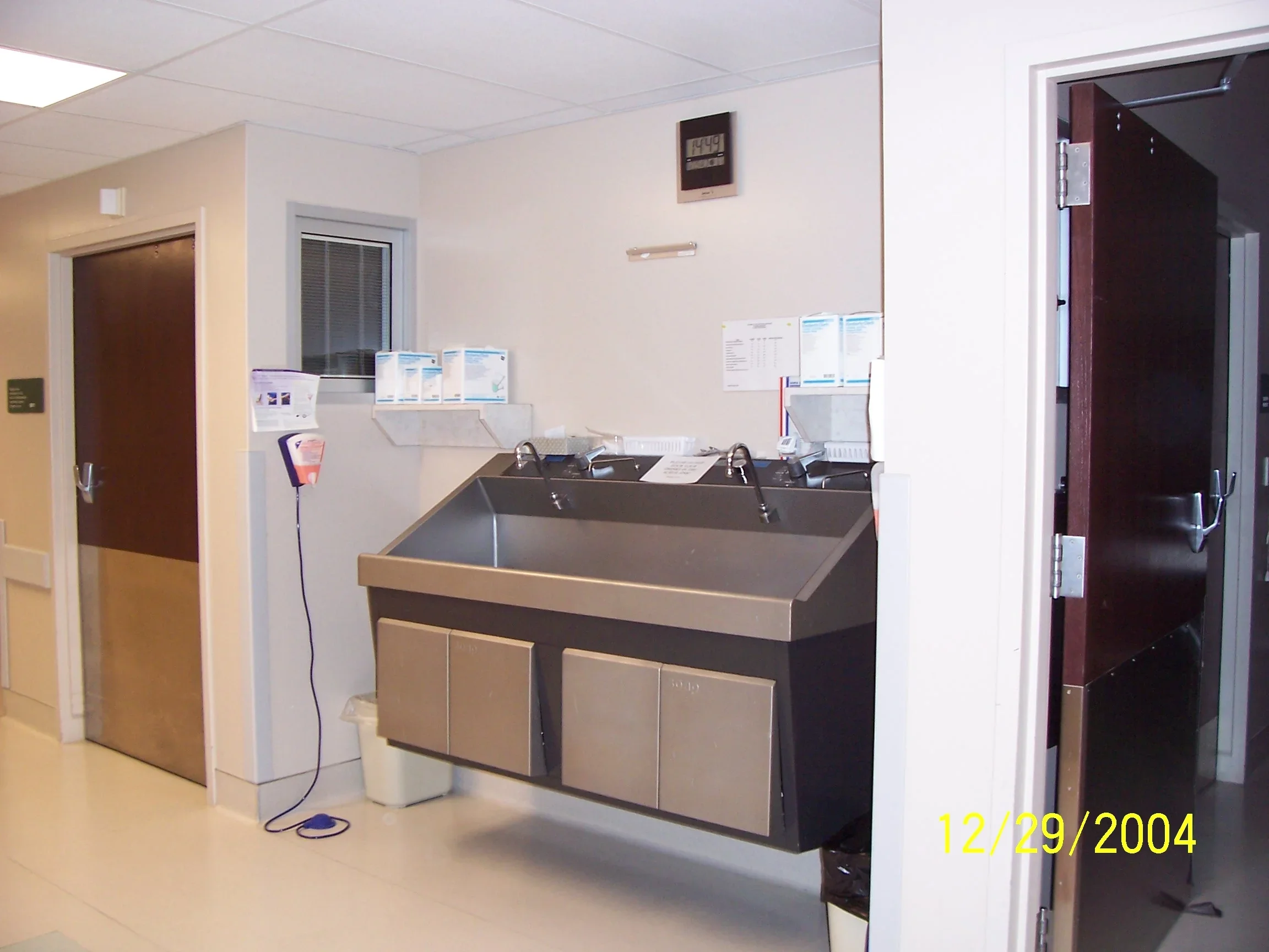 A hospital or medical facility's sink area with a stainless steel sink, paper towels, a digital clock displaying 12:49, and medical supplies on a counter, with a door and a window in the background.