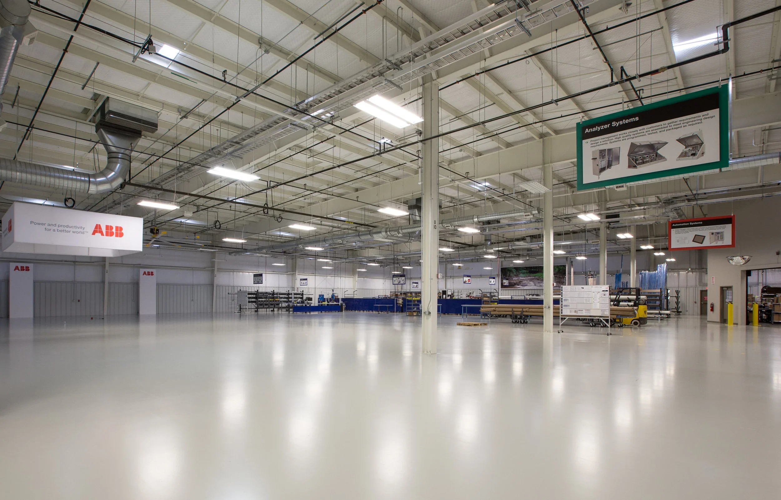 Empty industrial warehouse with white floors and high ceiling, displaying machinery, signs, and safety equipment.