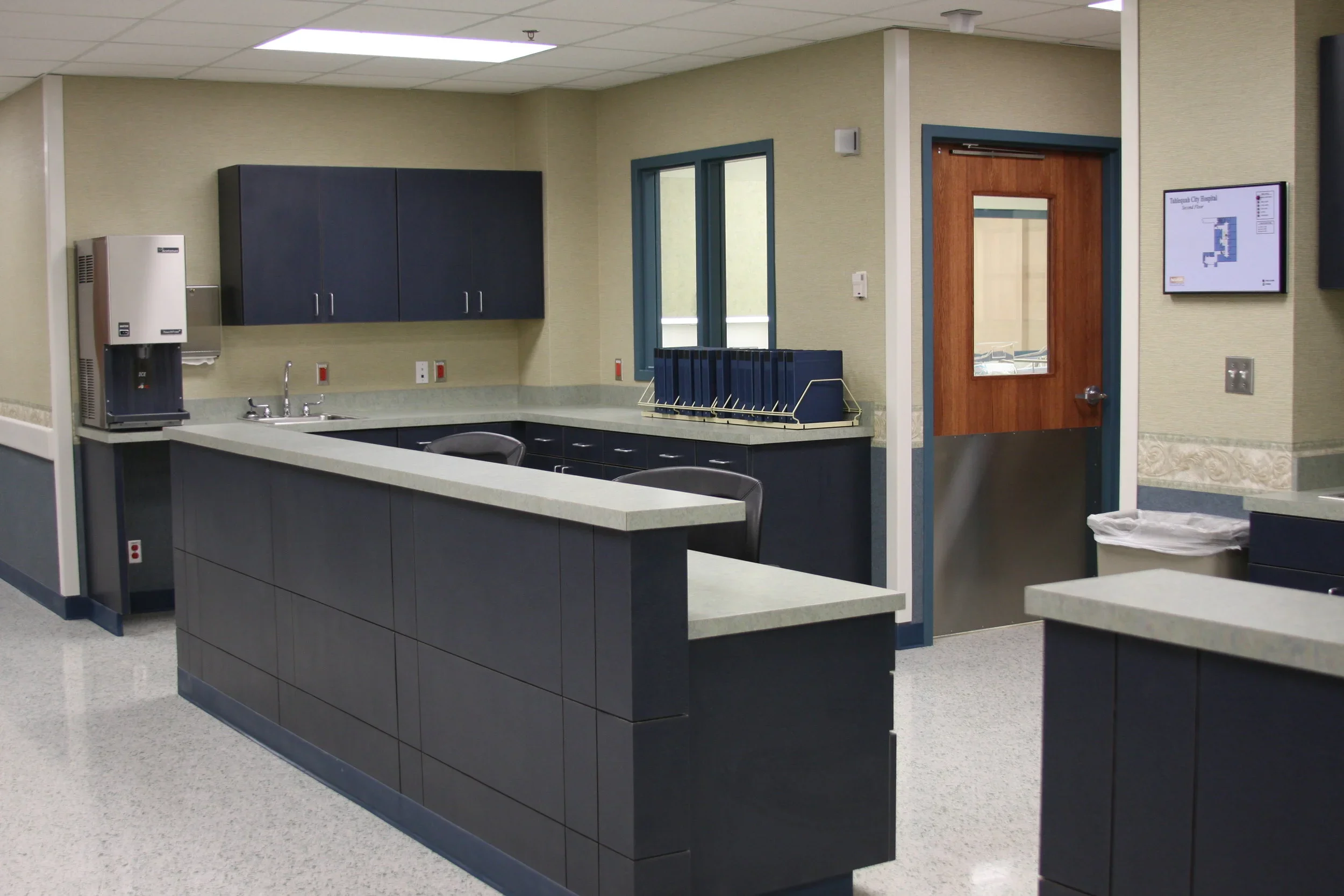 An empty break room or kitchenette with dark blue cabinets, a water dispenser, a sink, a window, and a wooden door with a window, in a professional or institutional setting.