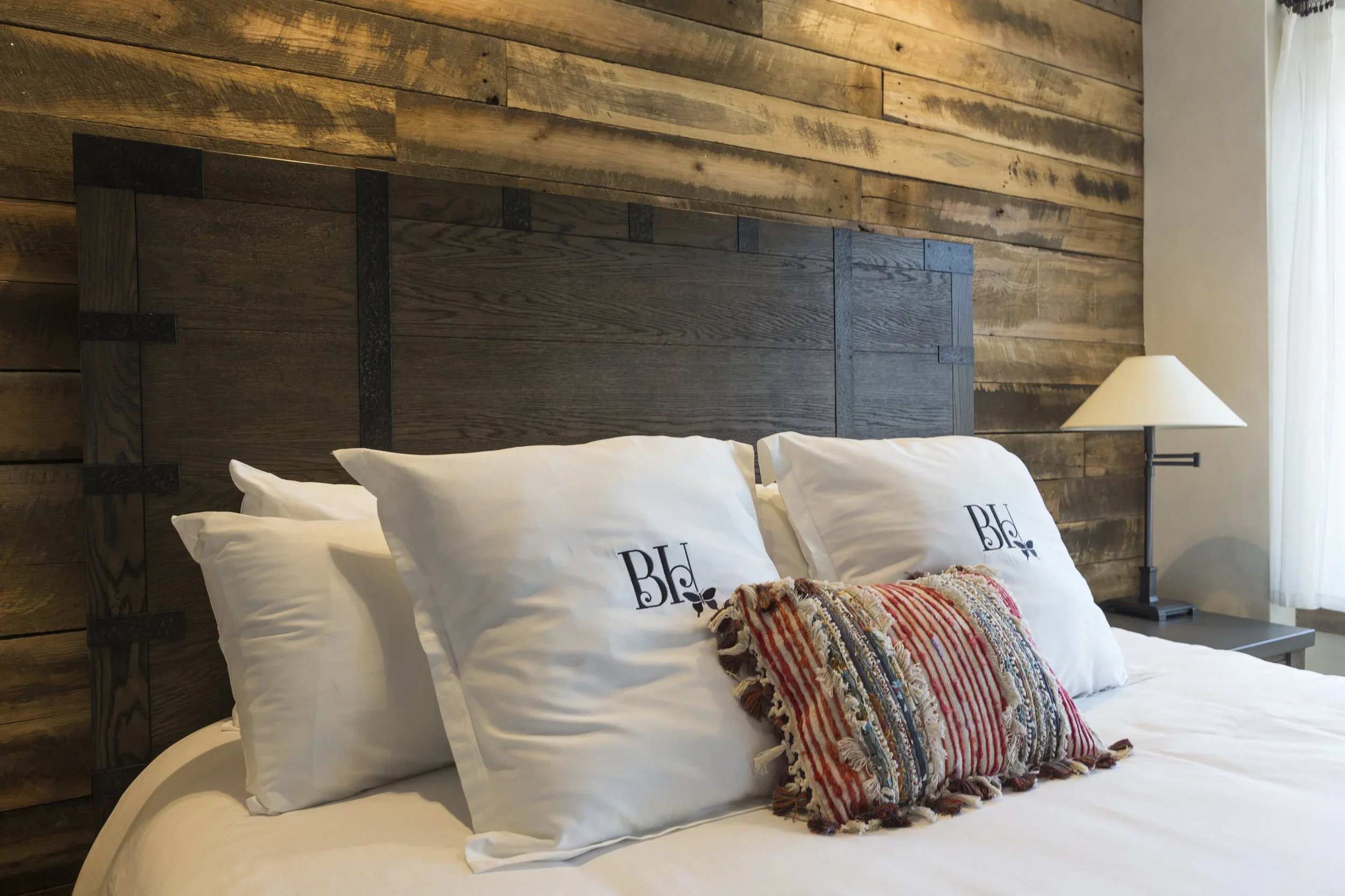 A neatly made bed with white pillows that have 'BE' written on them, a colorful decorative pillow, a dark wooden headboard, a wooden wall background, a side table, and a white lamp, in a cozy bedroom with a window.