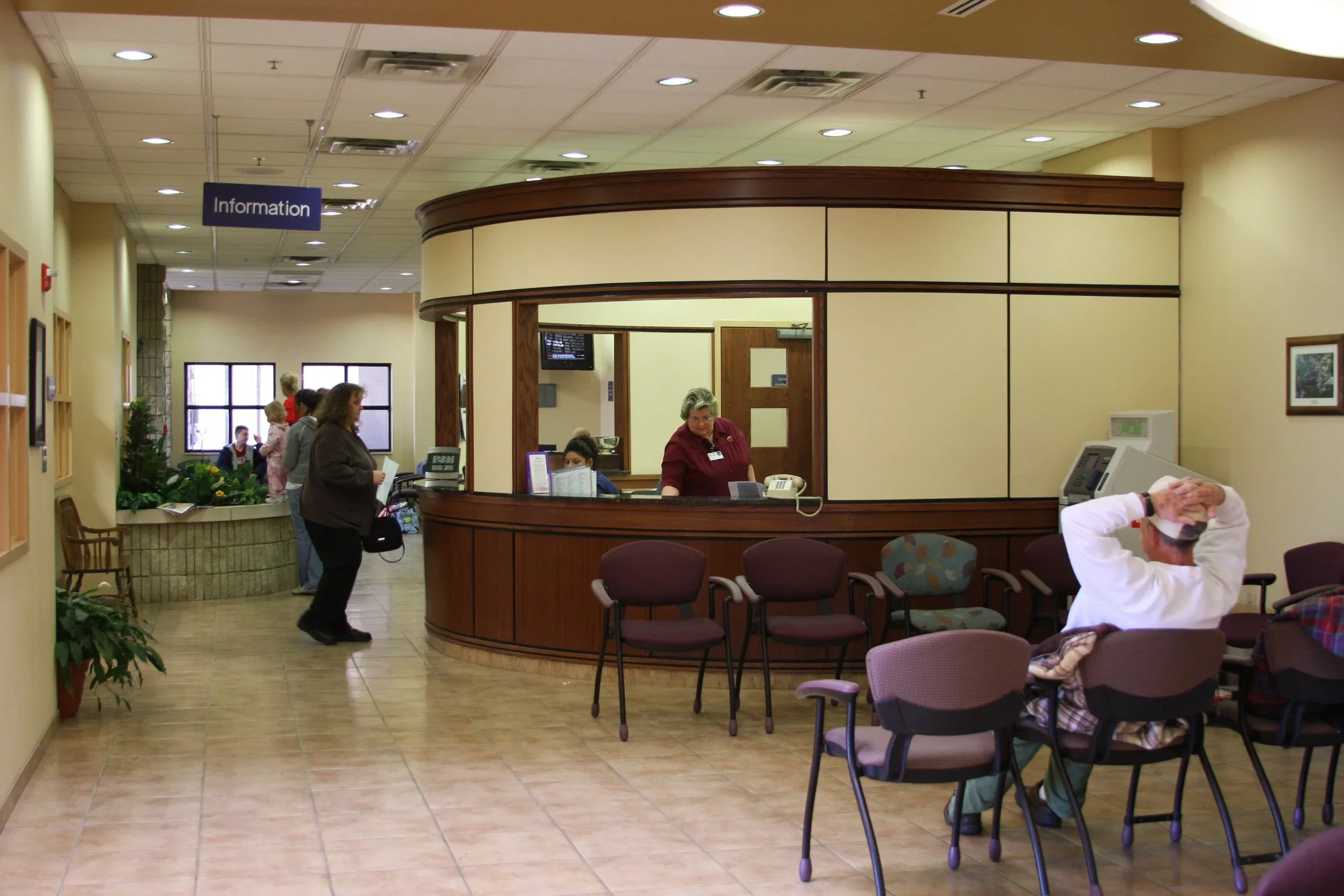 Hospital lobby with a reception desk, several chairs, and patients waiting; a woman at the desk and a man sitting with hands on head.