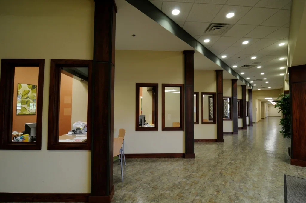 Empty hallway in an office or medical building with multiple rooms and large windows showing inside offices, decorated with green plants and artwork, with a low ceiling and bright lighting.