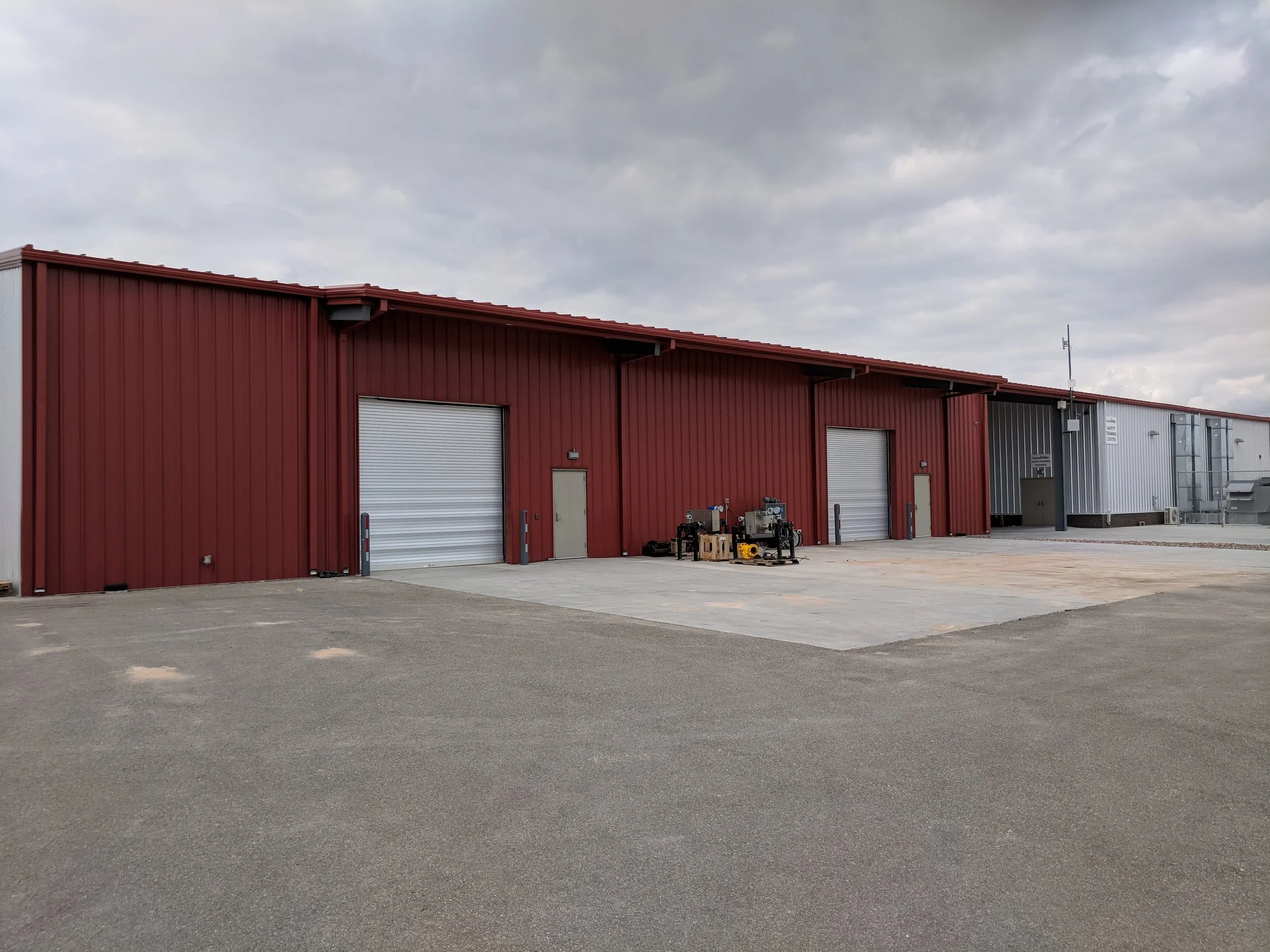 Industrial warehouse building with red and gray metal siding, multiple large roll-up doors, and a concrete parking area, under an overcast sky.