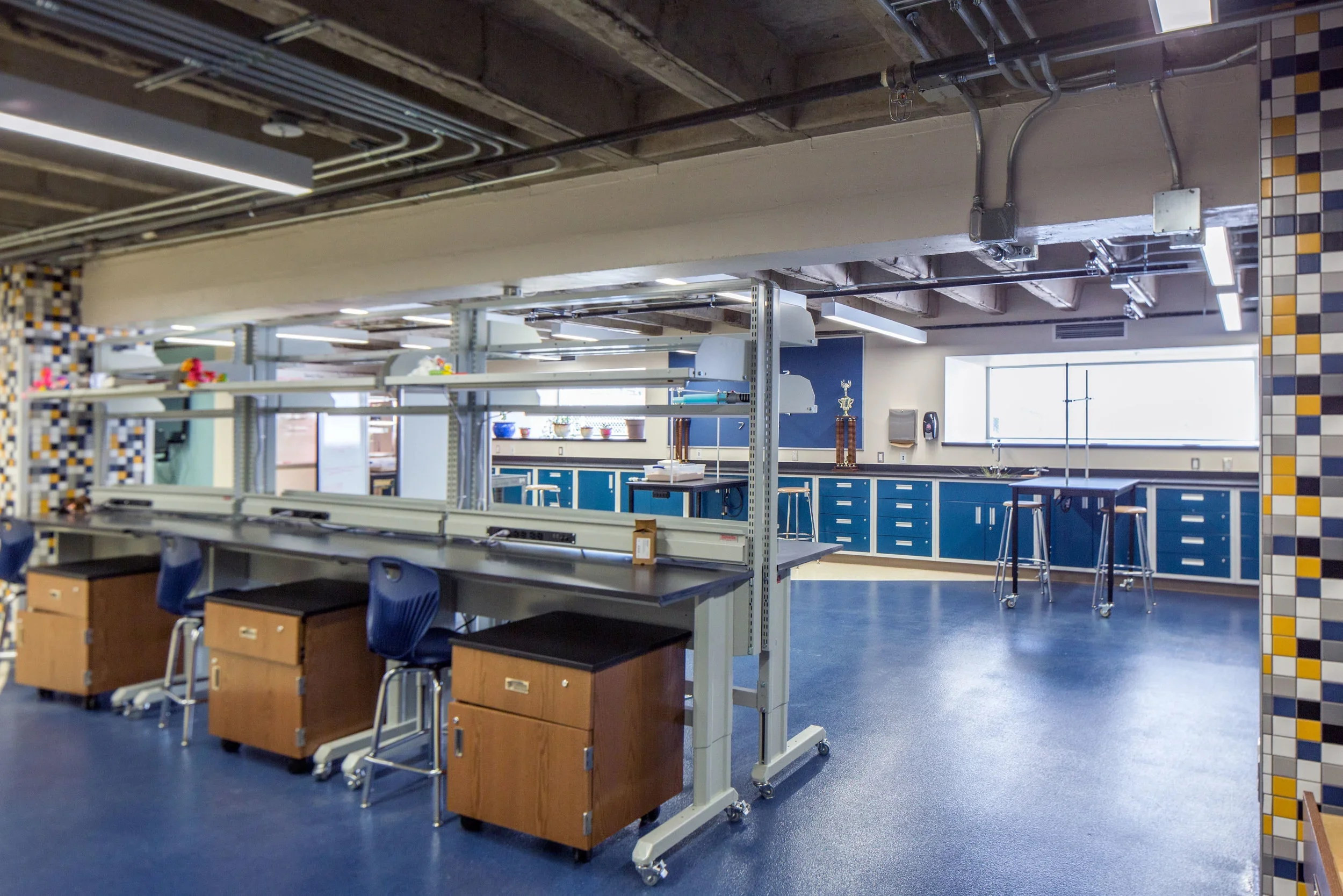 A spacious classroom or laboratory with blue cabinets, counters, and stools near a large window, with a row of workstations, and trophies on a shelf.