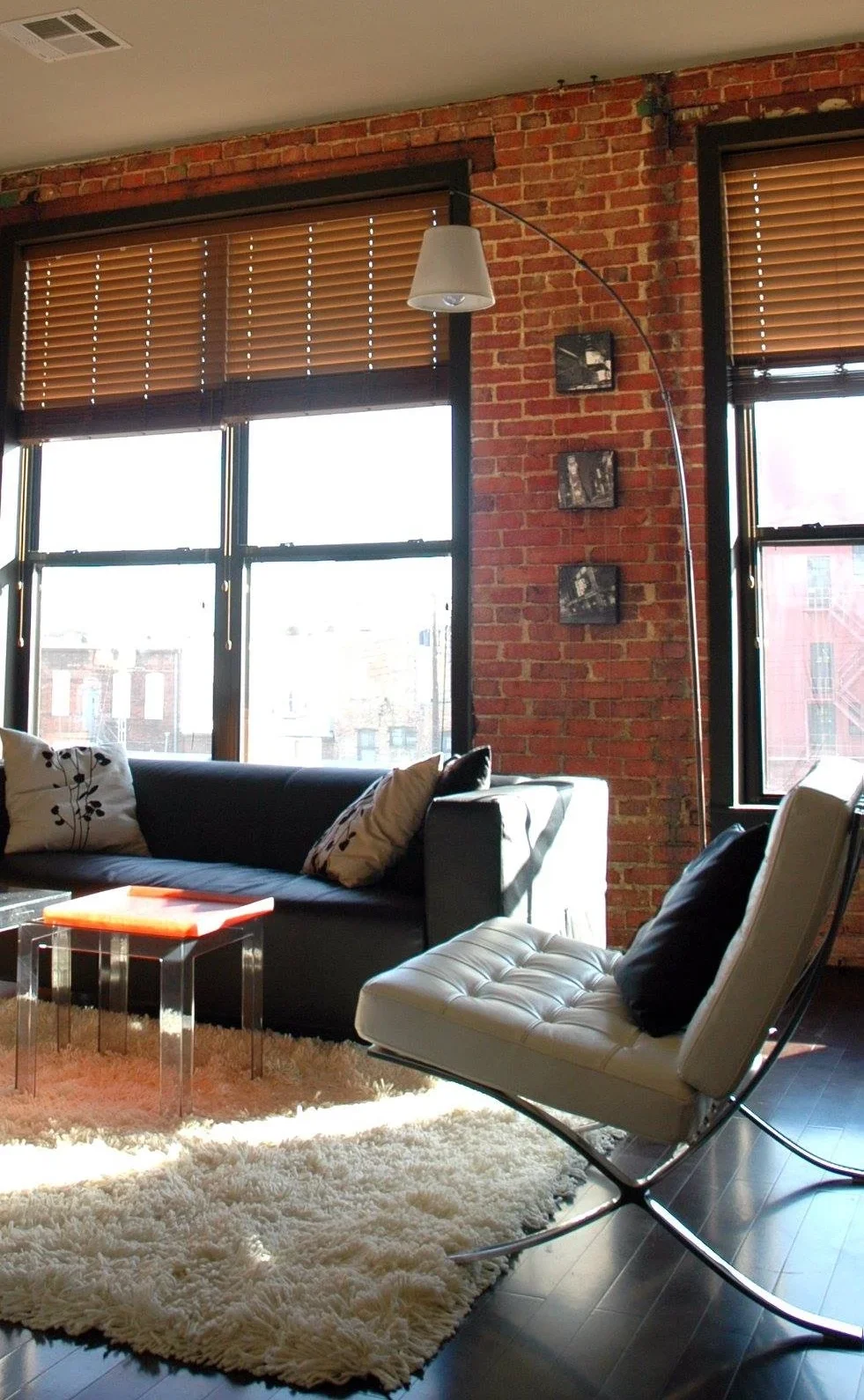A living room with exposed brick walls, large windows with wooden blinds, a black sofa with pillows, a white leather chair with a black pillow, a glass coffee table with an orange item on it, a fluffy area rug, and a white arc floor lamp.