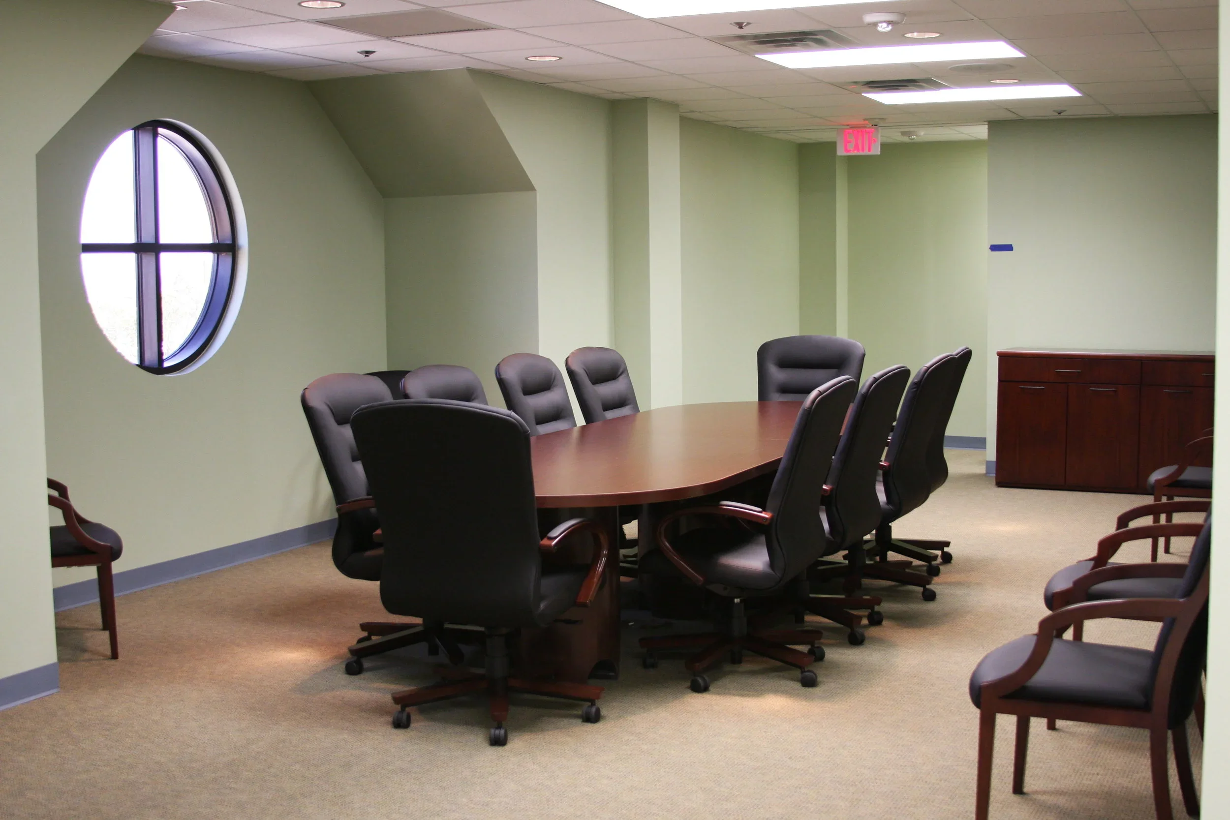 Empty conference room with a large oval table surrounded by black leather office chairs and additional chairs along the wall, beige carpet, green walls, round window, fluorescent ceiling lights, and a wooden cabinet in the corner.