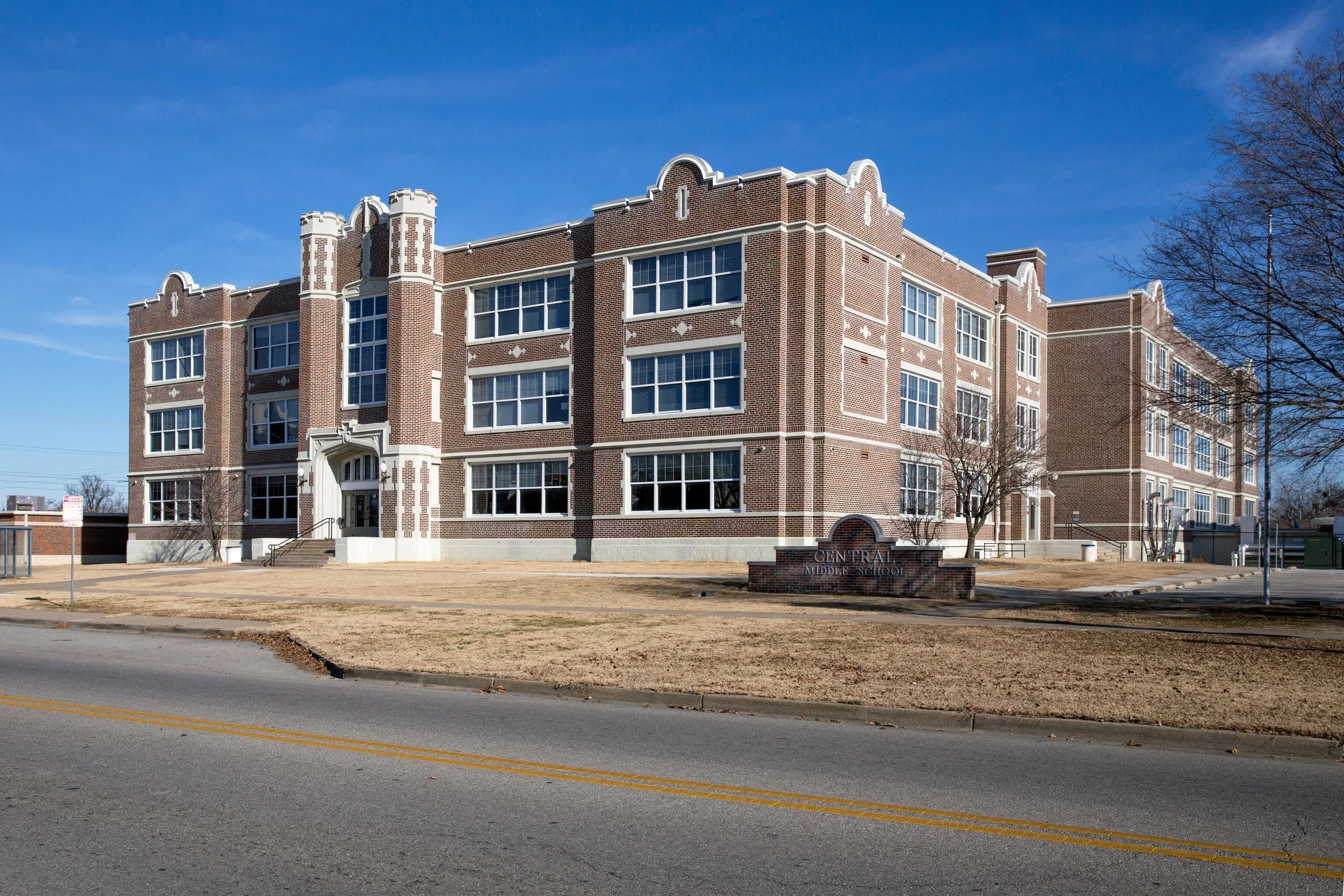 A large brick building labeled Central Middle School with white trim, multiple windows, and a front entrance with steps, on a clear day with a blue sky.