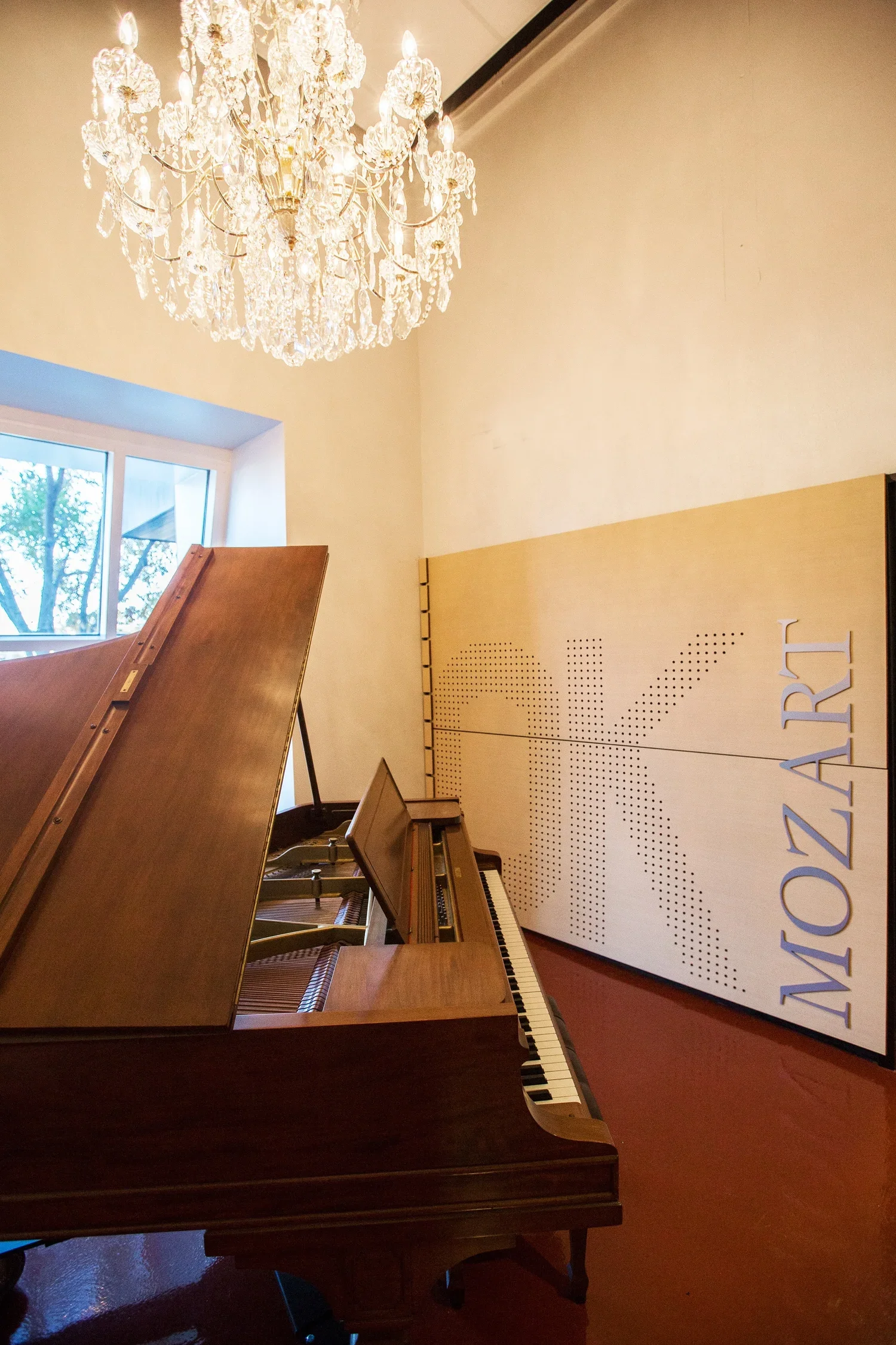 Interior of a room with a grand chandelier, a window with trees outside, a wooden grand piano, and a large wooden panel with 'OK MOZART' written on it.
