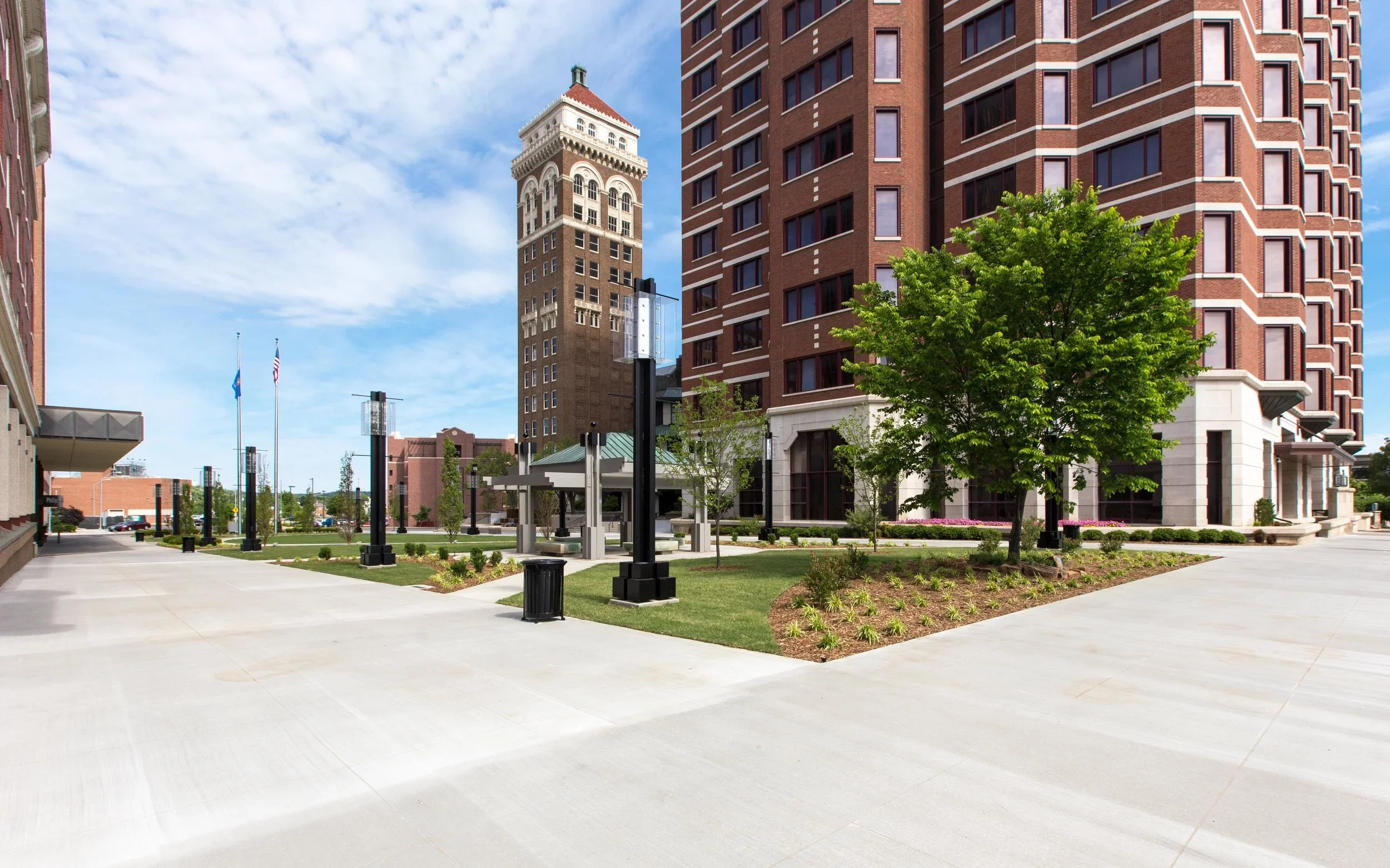 An urban plaza with a sidewalk, a large green tree, small shrubbery, black lampposts, and a small pavilion, surrounded by tall brick buildings, under a partly cloudy blue sky